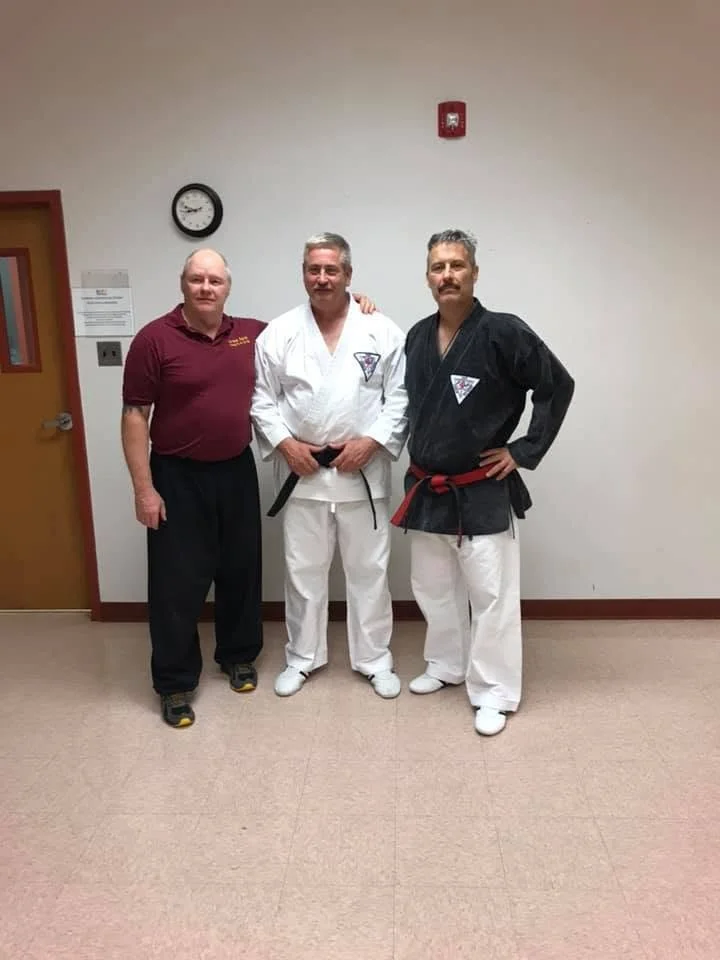 Three men standing together in a martial arts training room, two in martial arts uniforms and one in a maroon polo shirt, posing for a photo.