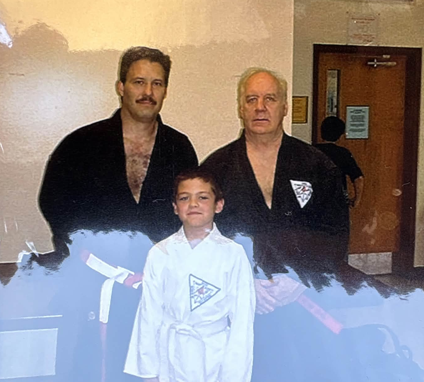 Three males, two adults and a child, dressed in martial arts uniforms, standing indoors in front of a beige wall and doorway.