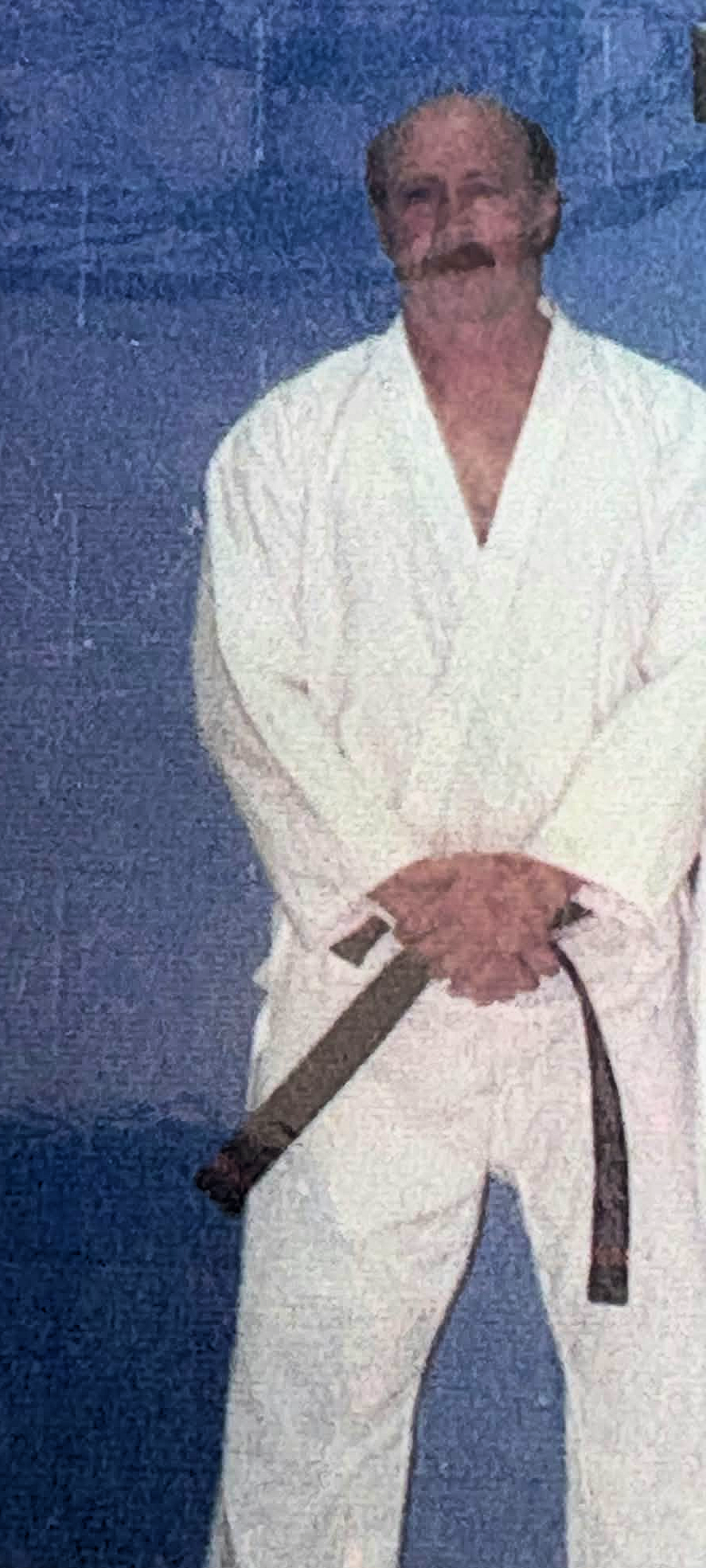 A man with a bald head, beard, and mustache dressed in a white gi holding a brown belt against a blue background.