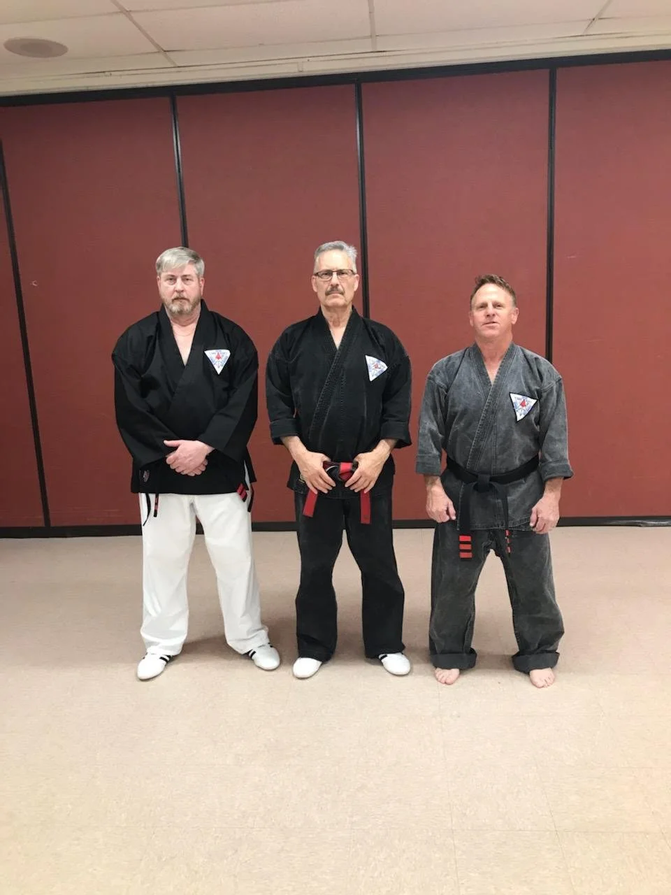 Three men in martial arts uniforms standing together in a training room with a plain red wall background.