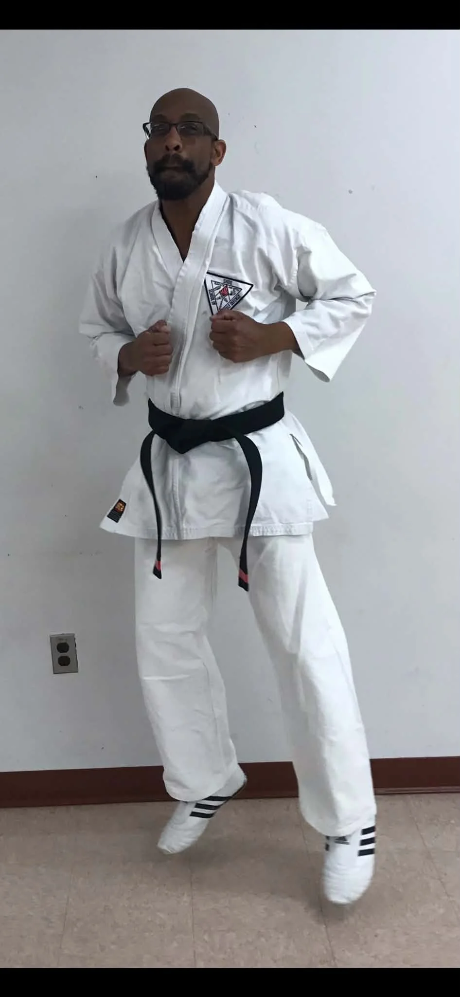 A man dressed in a white martial arts gi with a black belt and black-striped socks, standing in front of a plain white wall, posing with fists clenched and a confident expression.