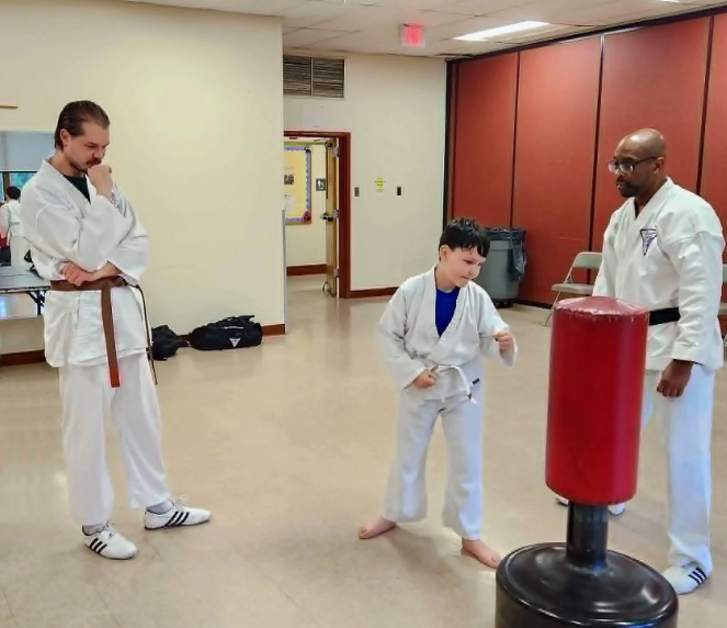 Three people in martial arts uniforms practicing with a red punching bag in a gym or dojo, with a young boy demonstrating a move while two instructors watch.