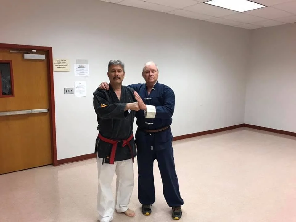 Two men in martial arts uniforms posing together in a sparsely decorated room, one with a black gi and red belt, the other in a blue gi, both standing close with their hands clasped.