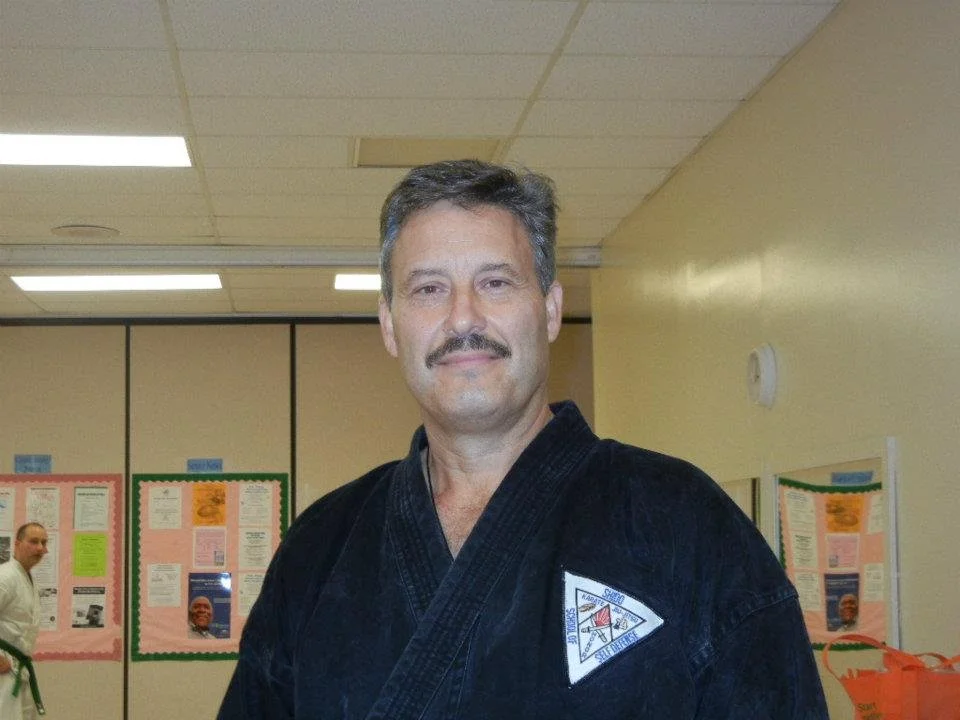 A man in a black martial arts uniform with a patch on the chest standing in a room with beige walls and bulletin boards, with another person in the background.