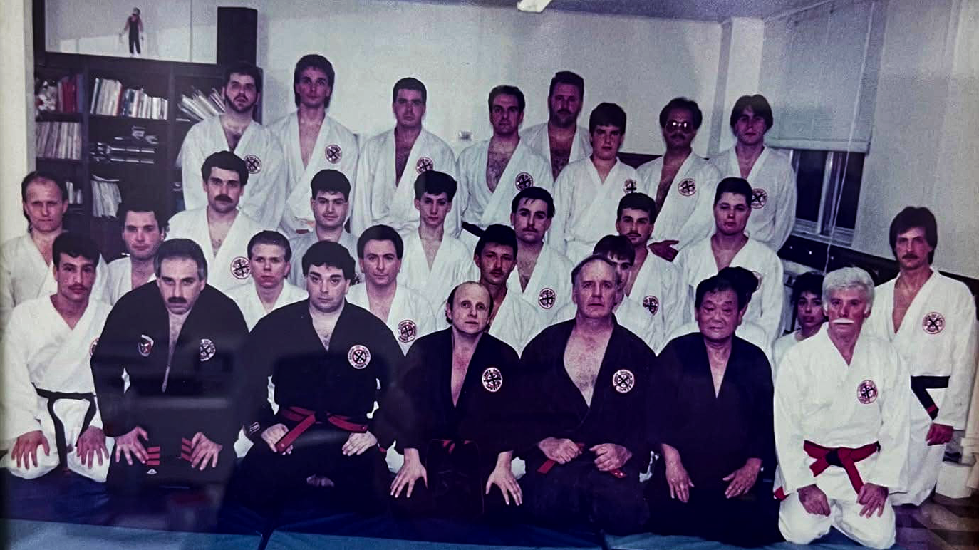 Group of martial artists in traditional uniforms with black and white belts, posing for a photo in a dojo or training room.