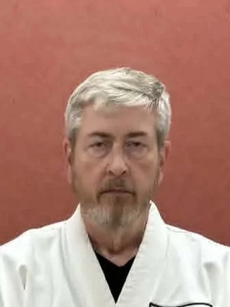A man with gray hair and beard wearing a white martial arts uniform, standing in front of a red wall.