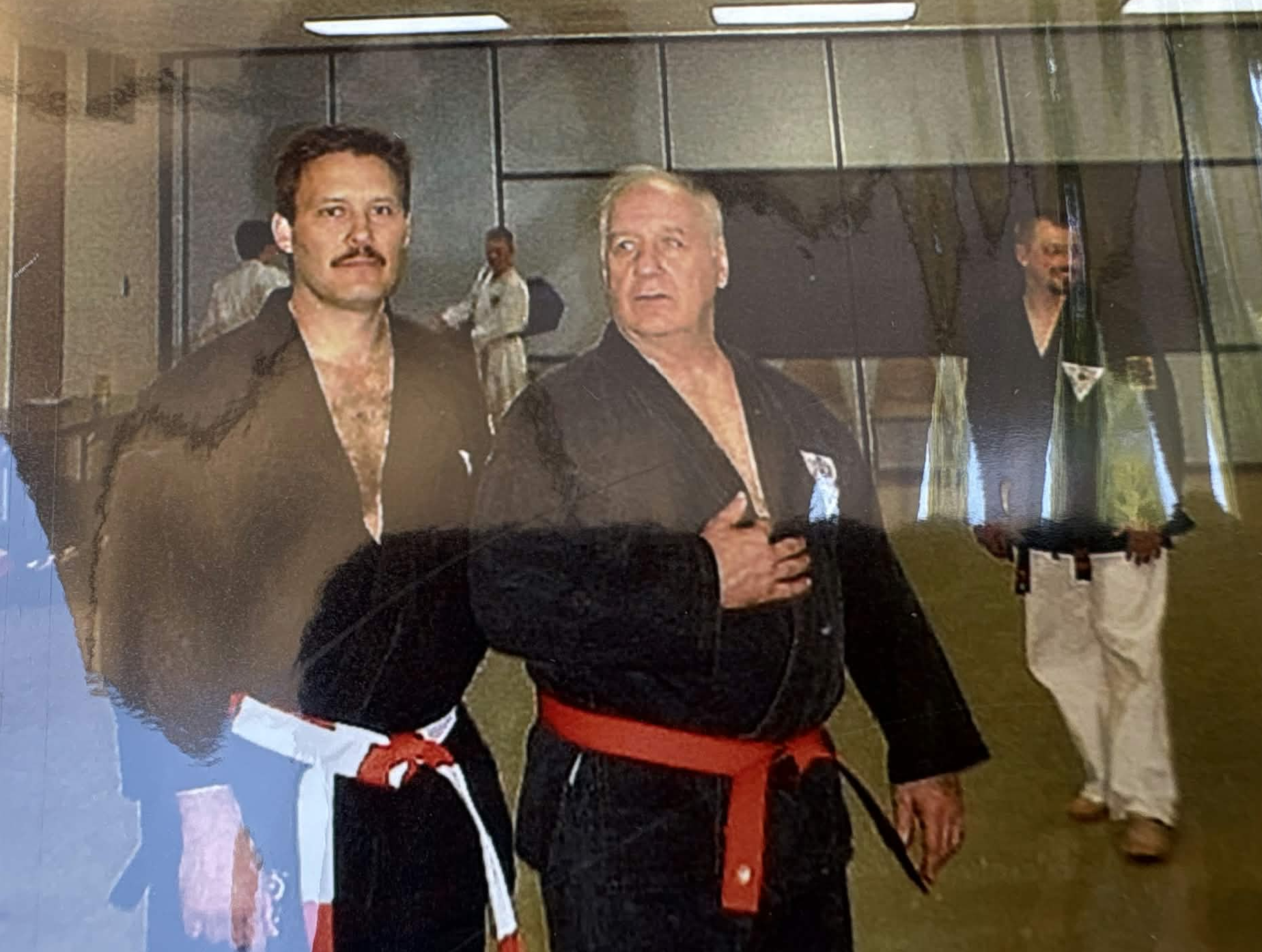 Two men in black traditional karate uniforms with red belts at a martial arts event, standing inside a building with other people in the background.