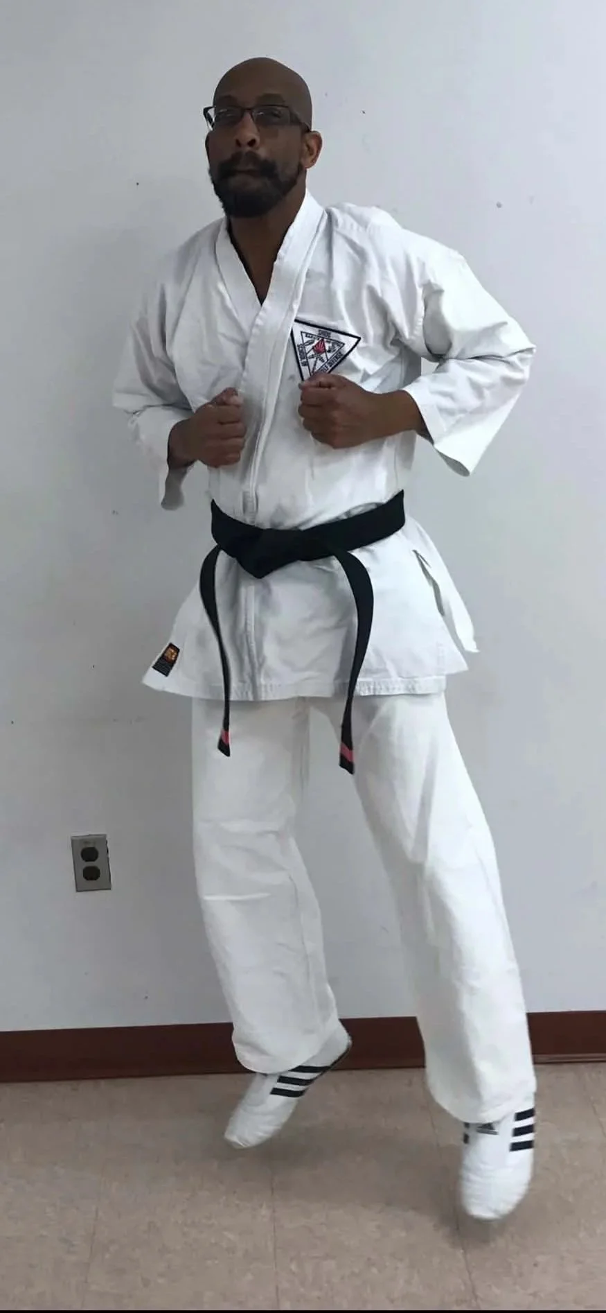A man dressed in a white martial arts uniform with a black belt, glasses, and a beard, standing in a martial arts stance against a plain white wall.