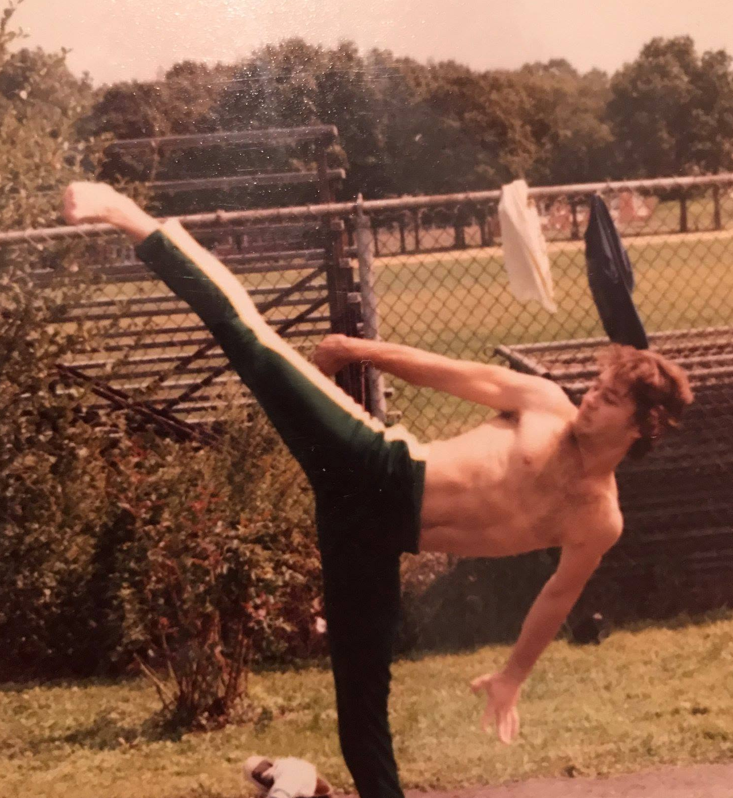 A young boy with curly brown hair doing a high kick outdoors, shirtless, wearing black pants, near a chain-link fence with clothes hanging on it, and trees in the background.