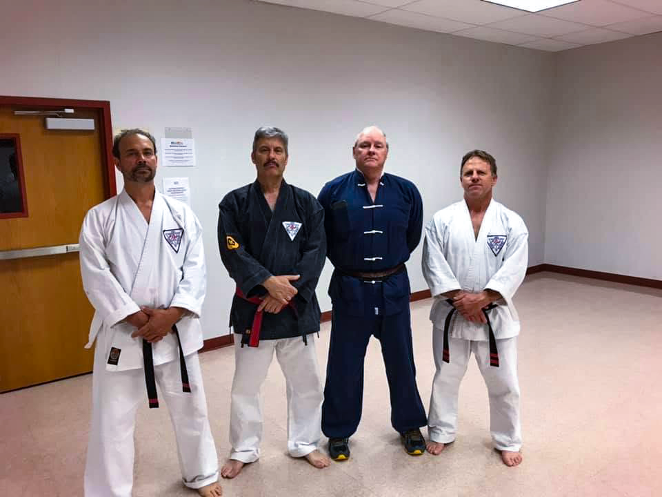 Four men in karate uniforms and one man in a martial arts gi stand in a room with a beige carpet and plain white walls.