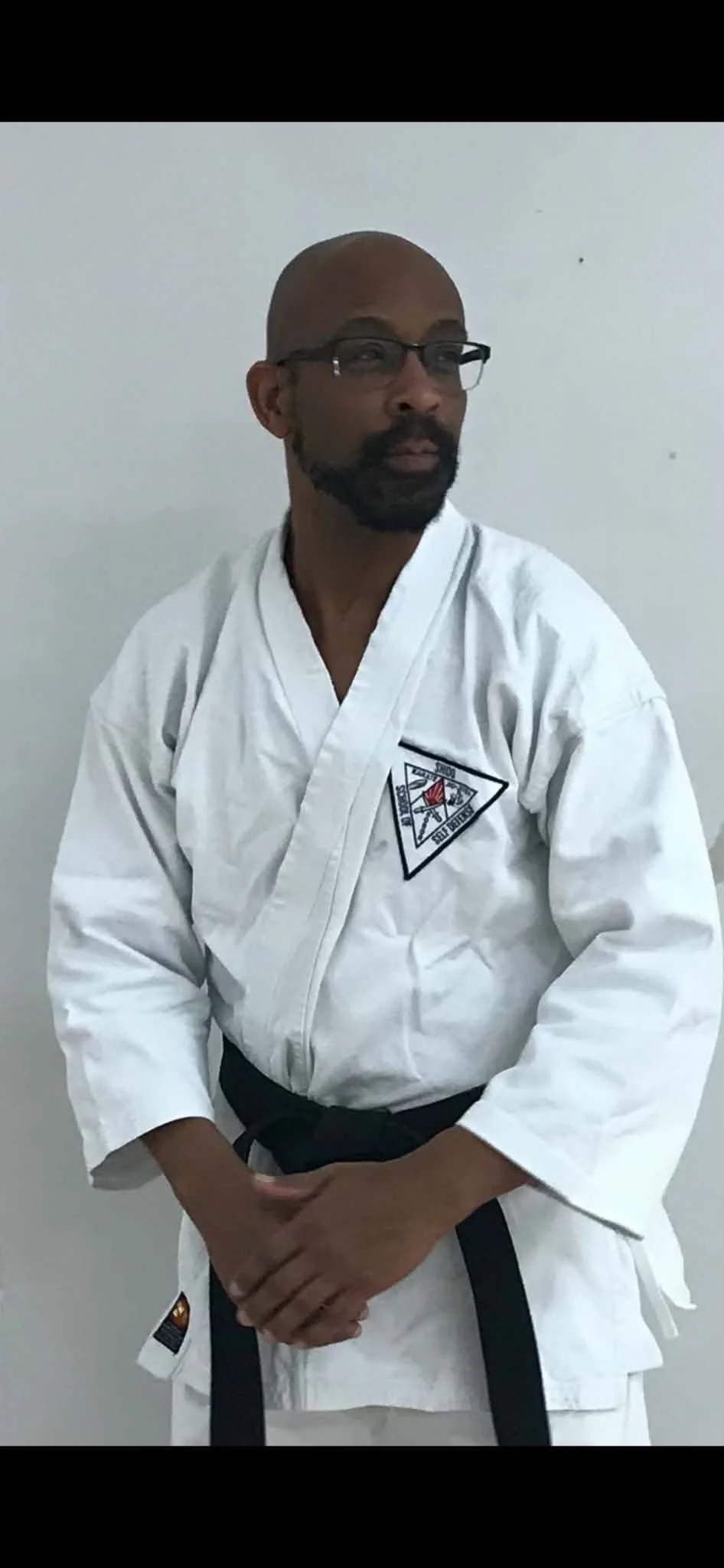 A man wearing glasses and a martial arts uniform with a black belt, standing against a plain wall, looking to the side.