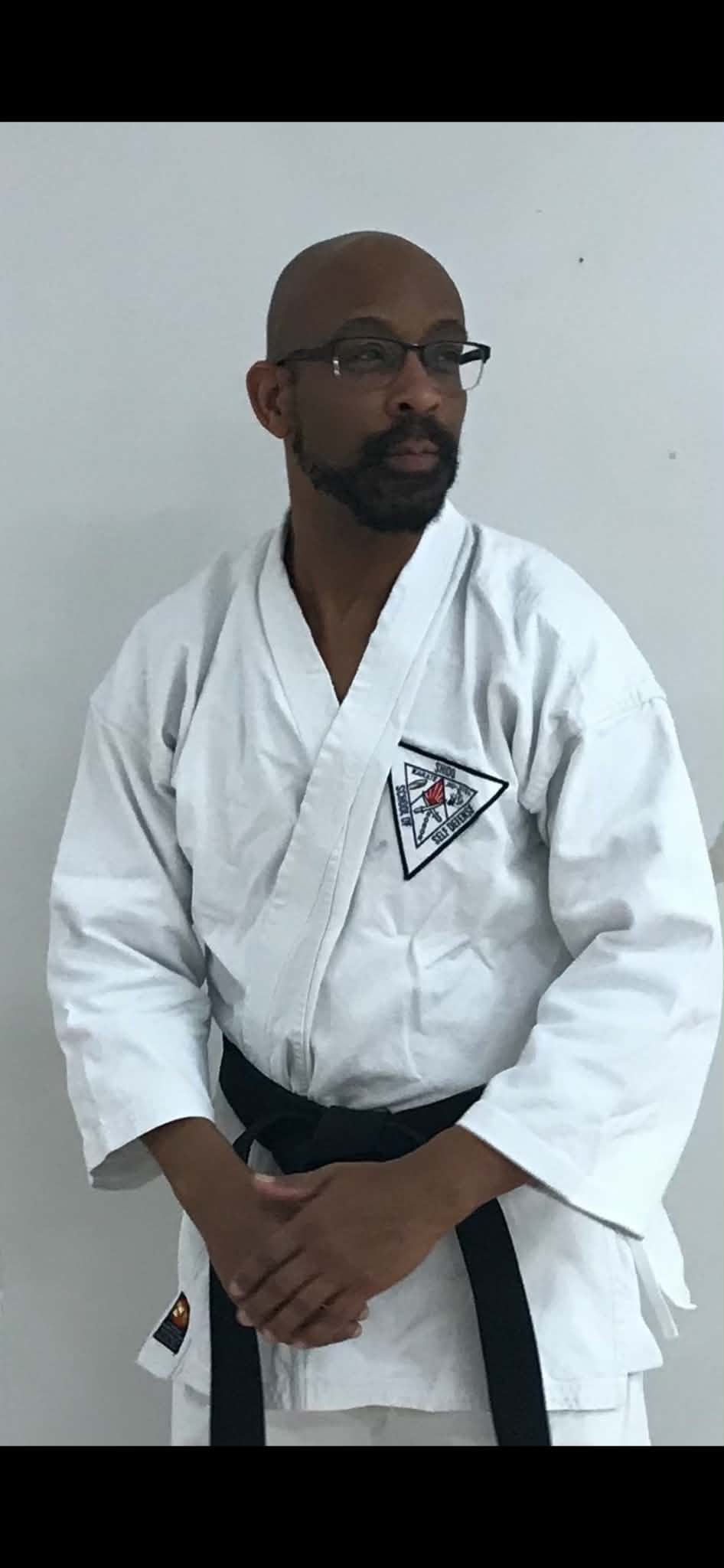 A man in a white martial arts gi with a black belt, standing against a plain wall. He is wearing glasses and has a beard, with his hands clasped in front of him.