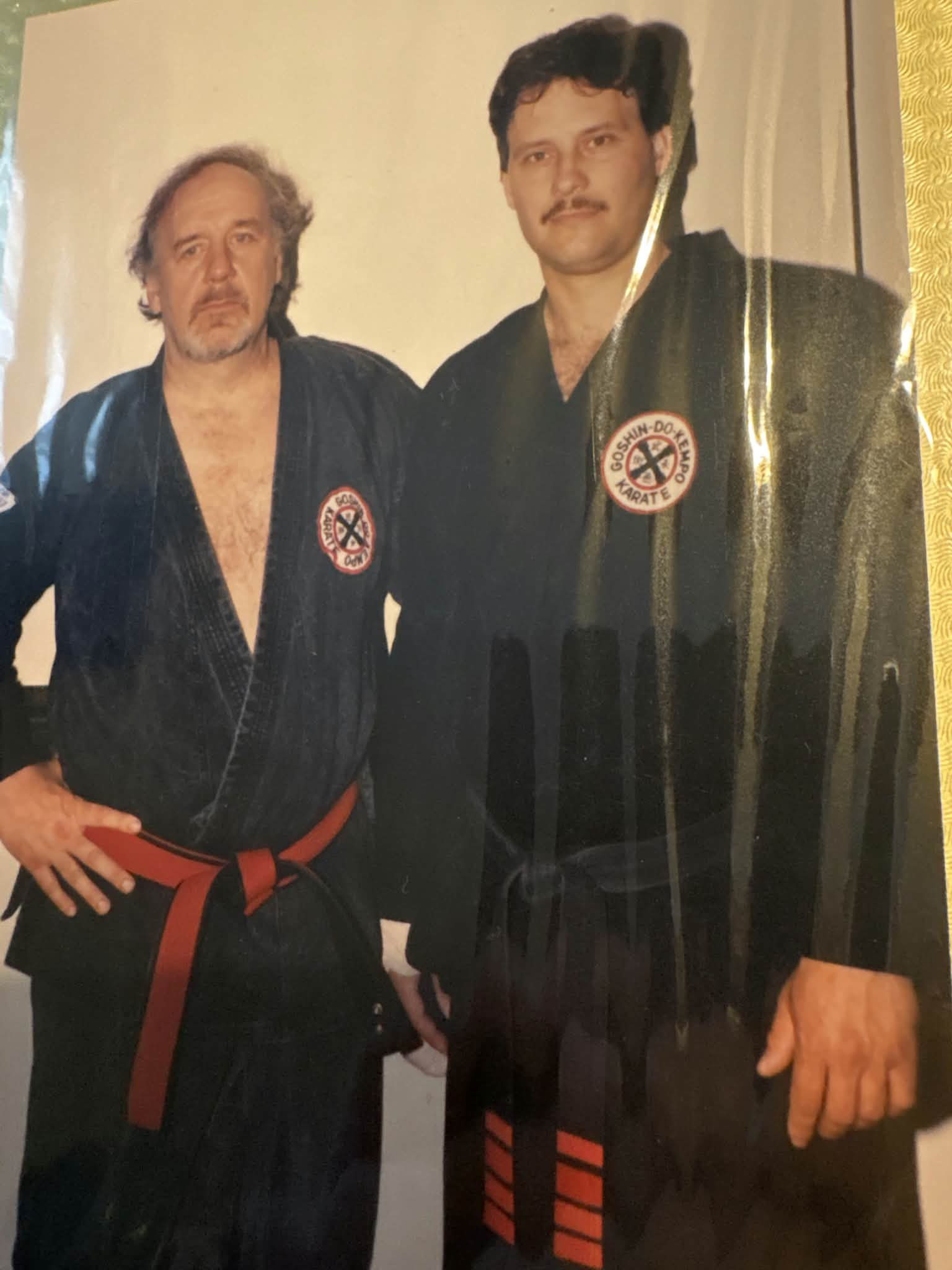 Two men wearing black martial arts uniforms with a patch that reads "GOSHIN-DO KENPO KARATE". One man has gray hair and a beard and is holding a wooden sword. The other man has black hair and a mustache.