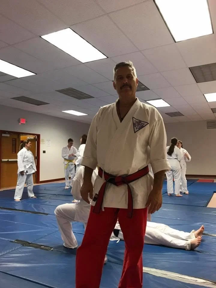 Martial arts instructor in a dojo, wearing a white gi with a black and red belt, standing on a blue mat with students practicing in the background.
