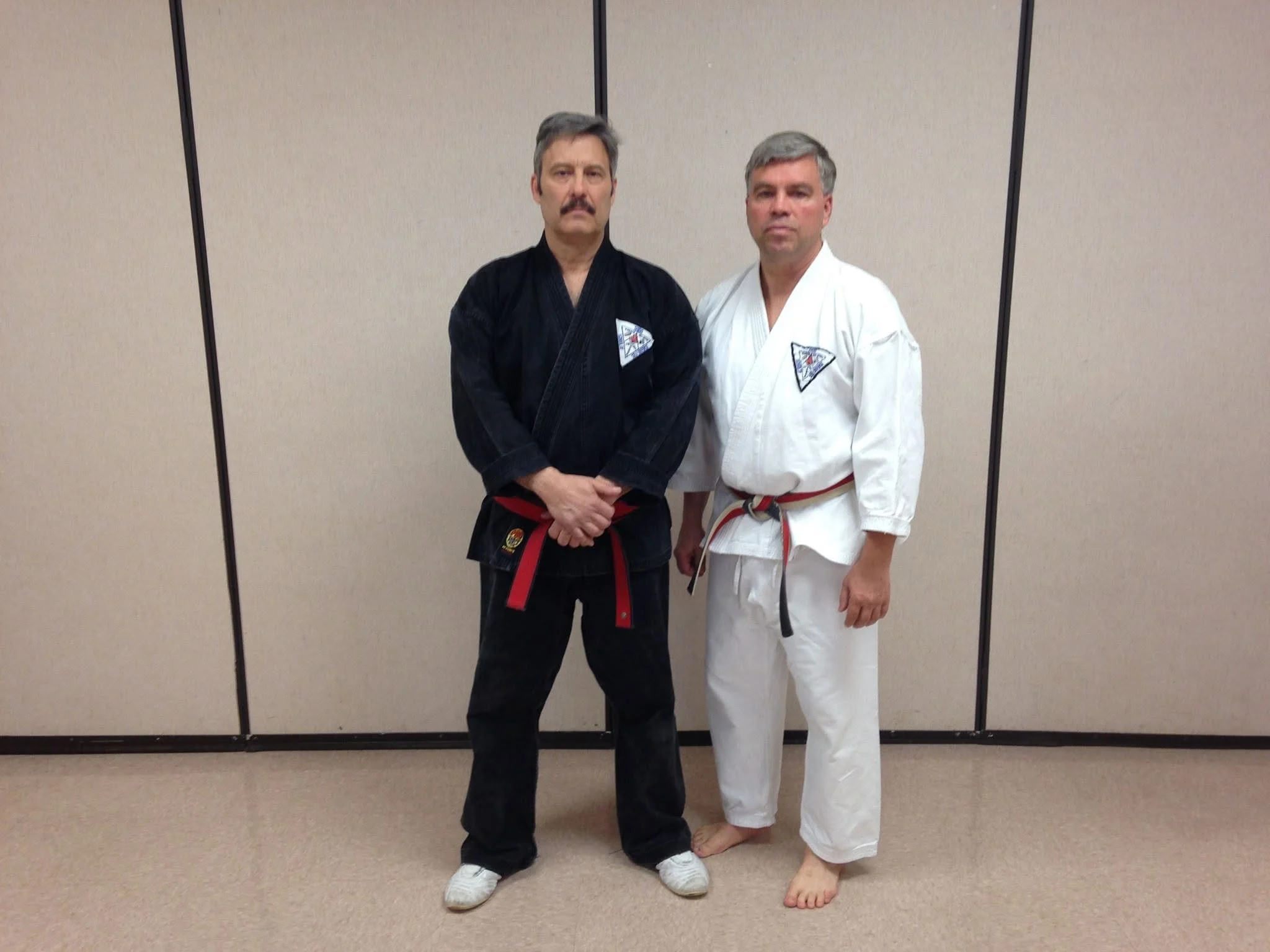 Two men in martial arts uniforms standing side by side in front of a beige partition. The man on the left wears a black gi with a red belt, and the man on the right wears a white gi with a red and white belt. They are in a martial arts training or competition setting.