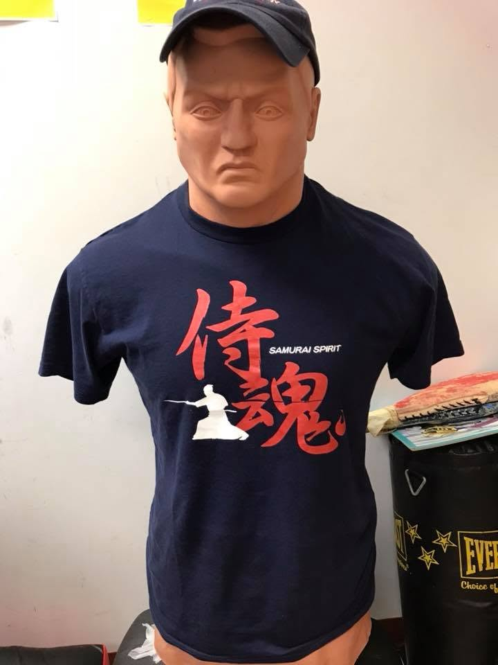 Mannequin wearing a black T-shirt with red Japanese kanji, white silhouette of a samurai, and the words 'SAMURAI SPIRIT' on it, in a room with white walls.