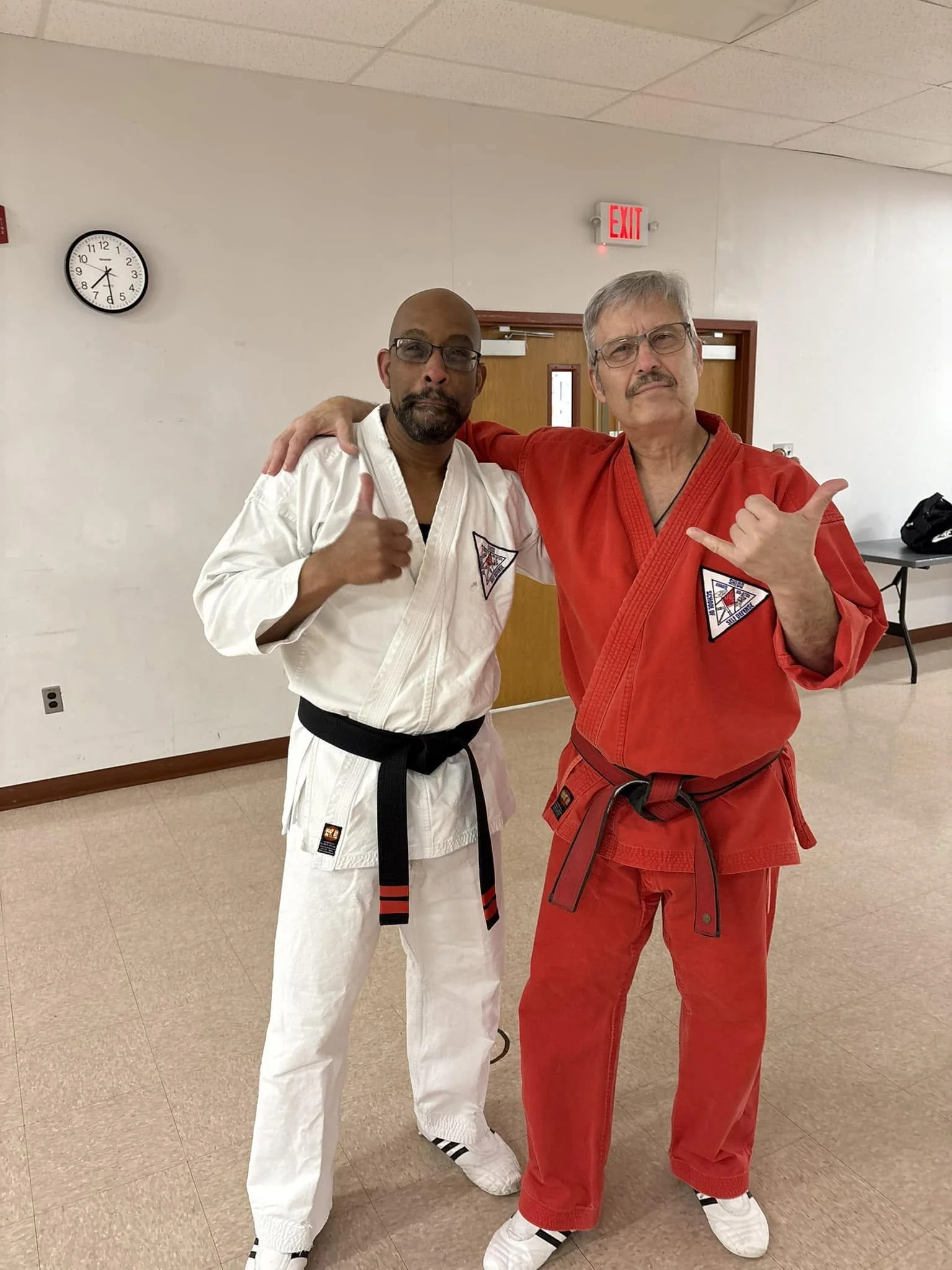 Two men in martial arts uniforms pose together in a room with beige walls and a clock on the wall showing 11:40.