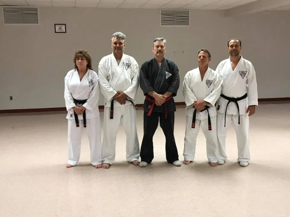 Five martial artists in karate uniforms standing in a dojo. The person in the middle wears a black gi with a red belt, and the others wear white gis with black belts. They are posing in a line with a beige wall behind them.
