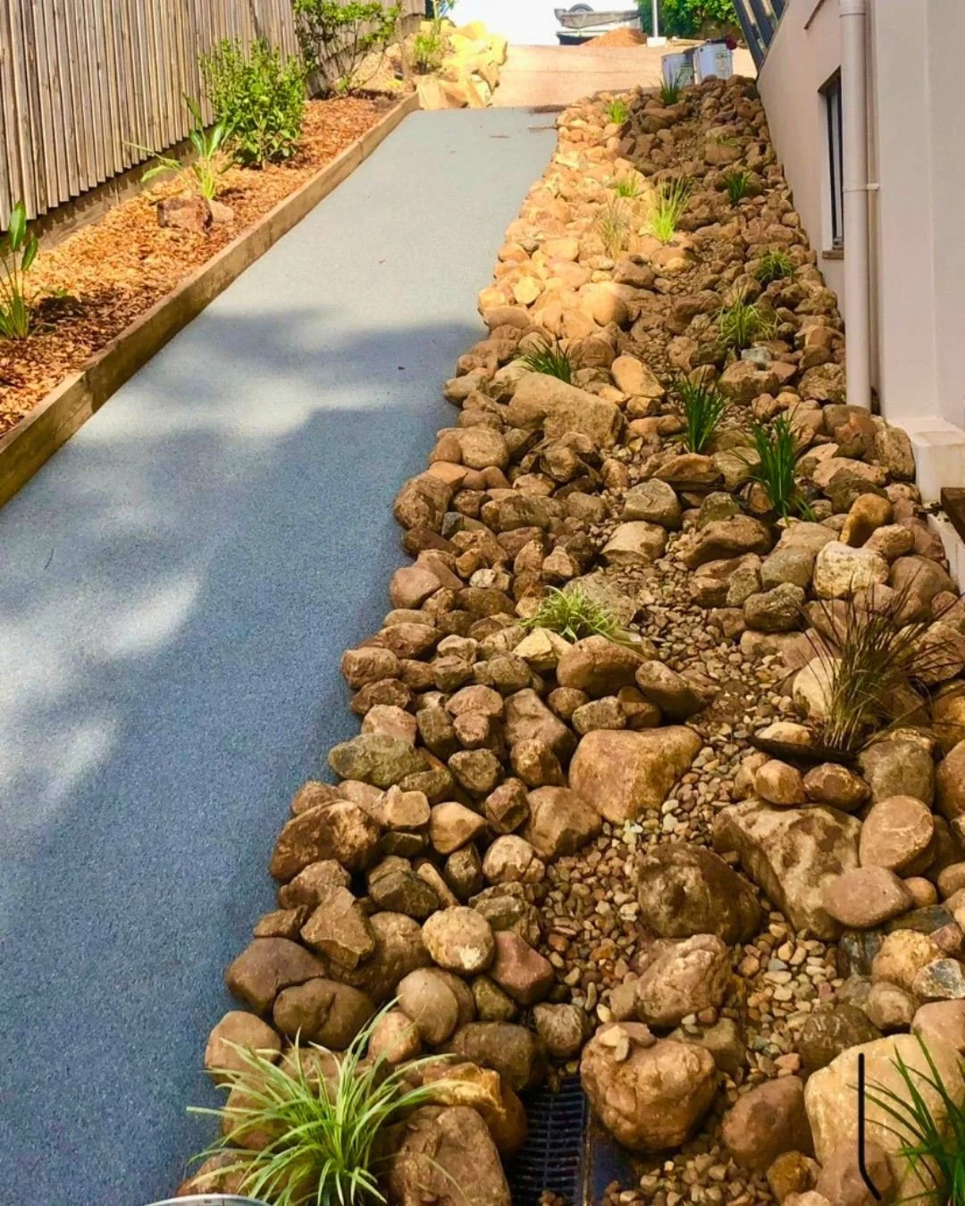 A pathway with a blue surface runs alongside a garden with rocks and small plants, with a wooden fence on one side and a white wall on the other.