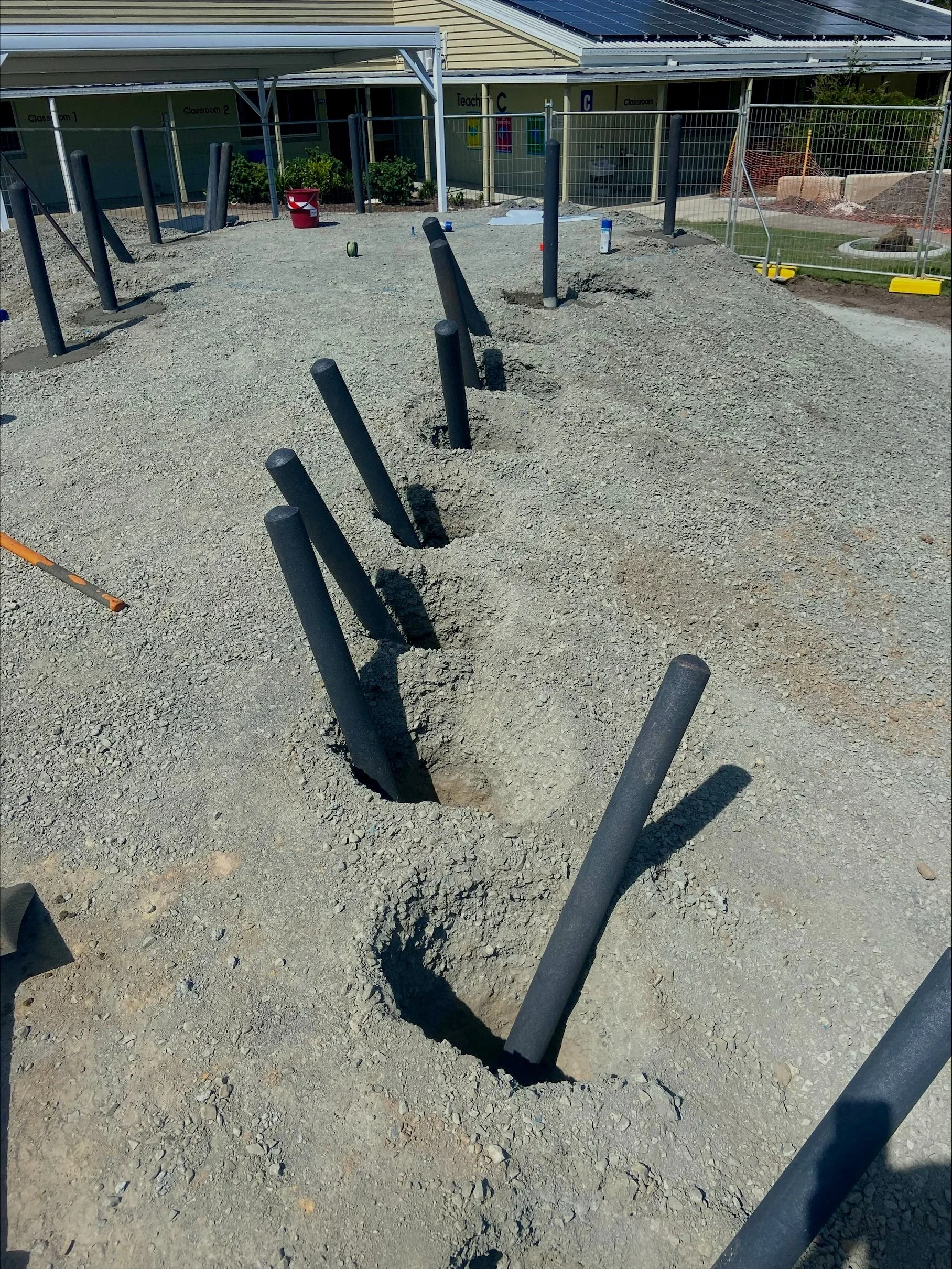 Construction site with large holes and black pipes sticking out of the ground, surrounded by dirt and gravel, with a fenced area and a building in the background.