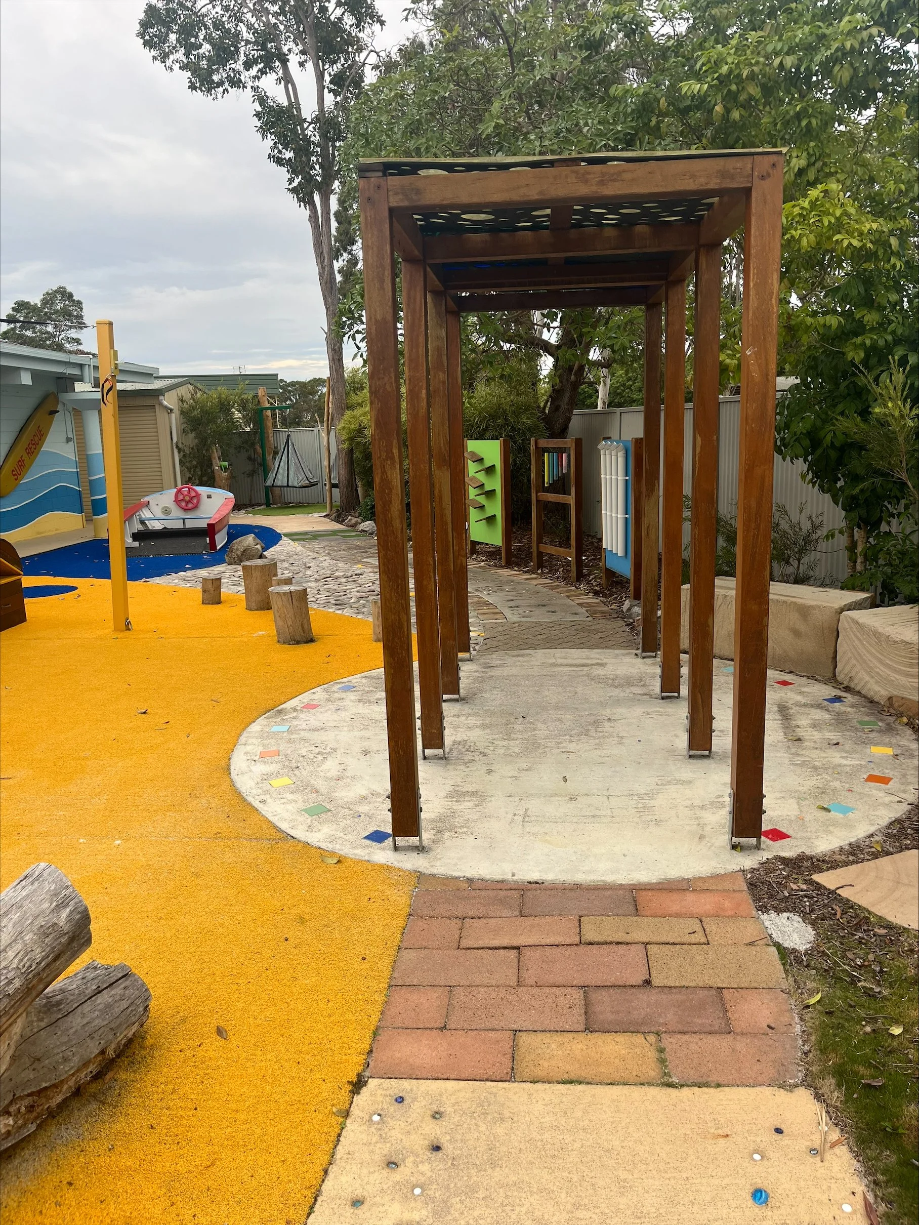 Colourful outdoor play area rubber surfacing, a boat-shaped sandbox with a pink steering wheel, wooden posts forming a pathway or arch, playing panels, and a pathway with multicolored tiles, surrounded by trees and a metal fence.