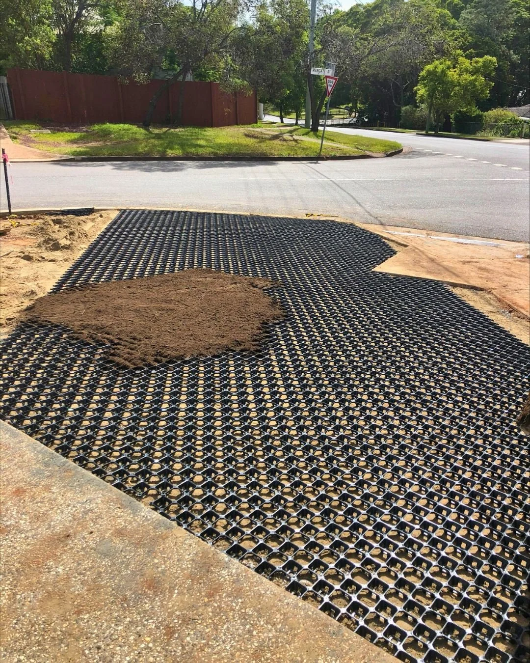 A section of sidewalk under construction with plastic grid base and a pile of dirt on top, adjacent to a street intersection.
