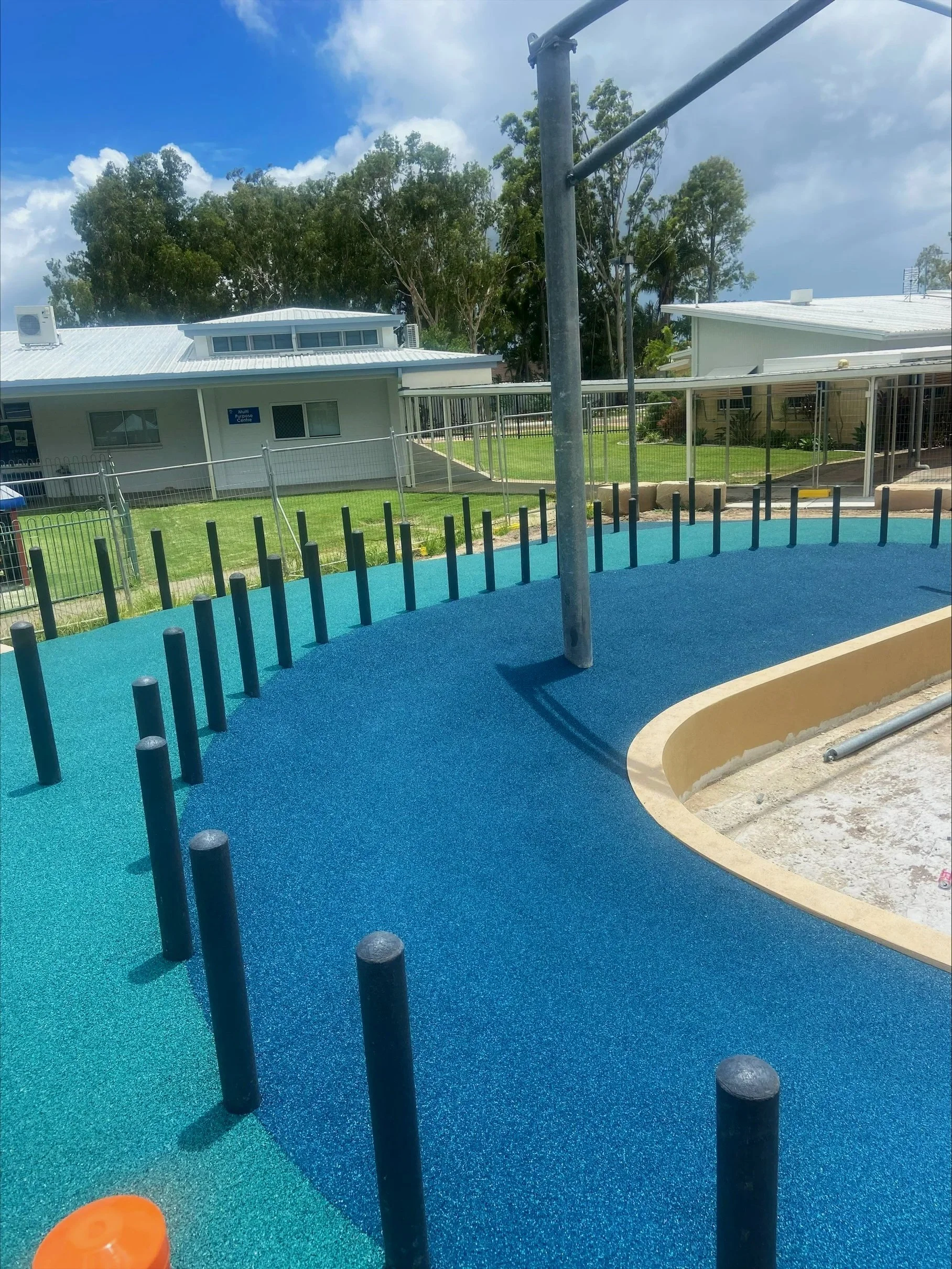 Playground with blue rubber flooring, black posts, and metal pole, grassy area, white building with a sign, trees and cloudy sky in the background.