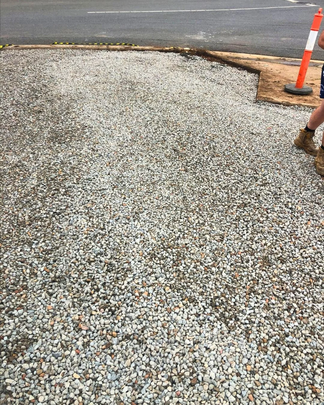 A gravel area being prepared for paving, with some construction barriers and a worker's legs visible.