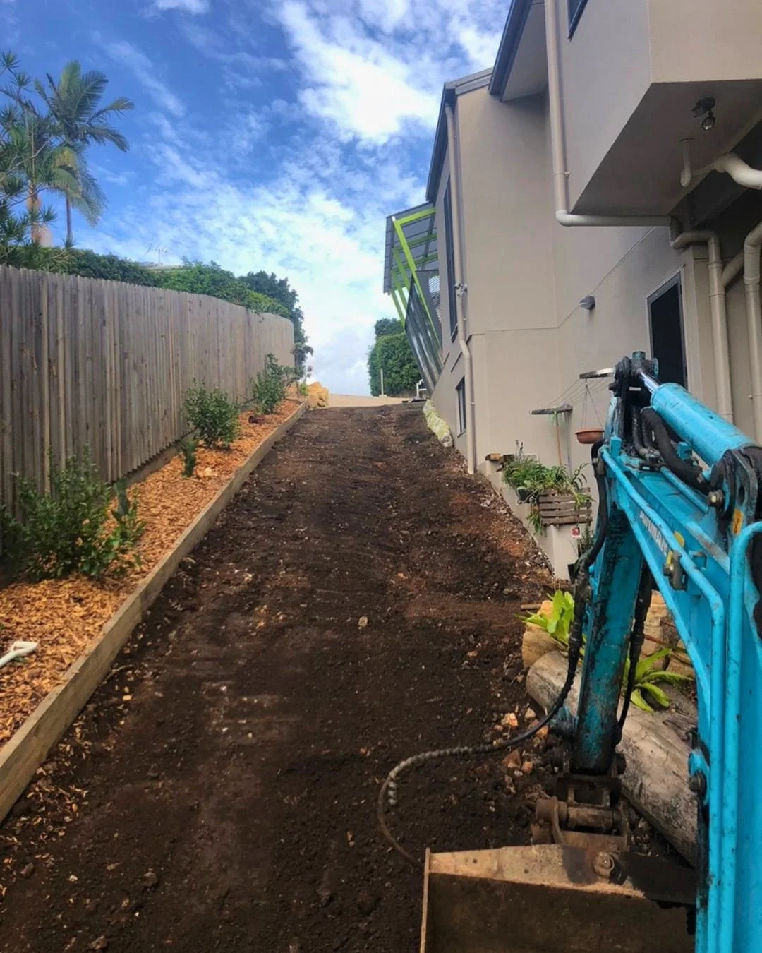 A sloped backyard with freshly tilled dark soil, a wooden retaining wall with small plants on the left, a house with piping on the right, and a blue compact excavator in the foreground. A clear blue sky with scattered clouds overhead.