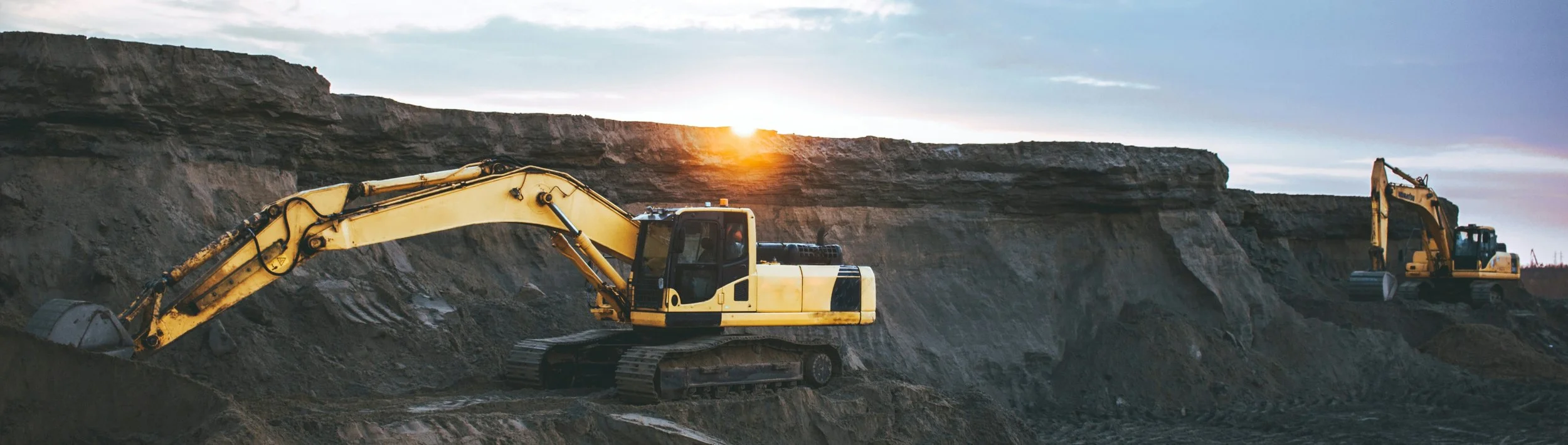 Dos excavadoras amarillas trabajando en un sitio de construcción con una montaña de tierra al atardecer.