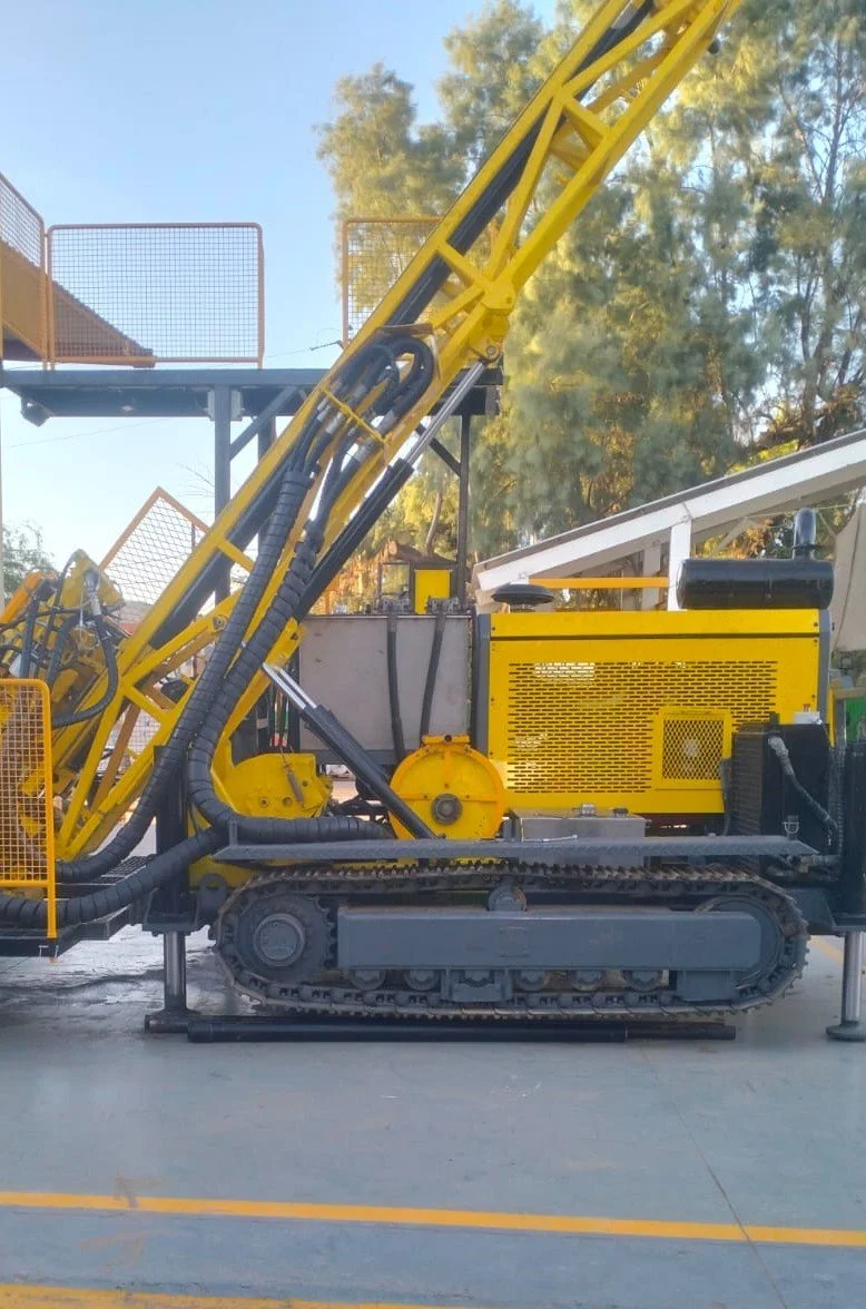 Maquinaria de perforación de color amarillo y negro en una superficie pavimentada, con árboles y cielo claro de fondo.