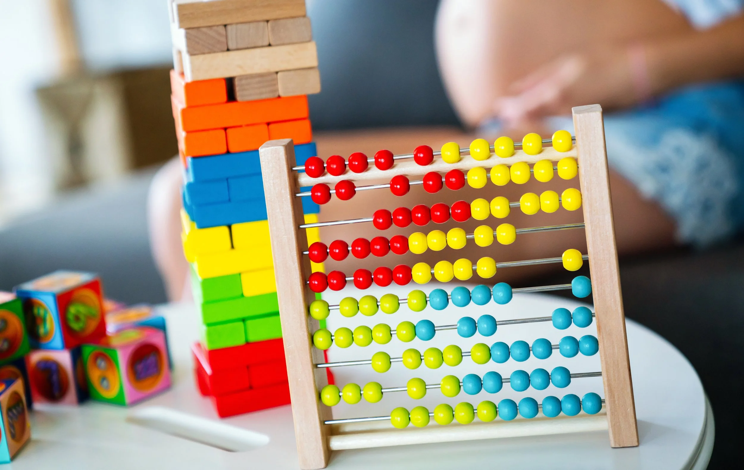 A close-up of a colorful wooden abacus with yellow, red, and blue beads, and a stacking block tower in the background on a white table.