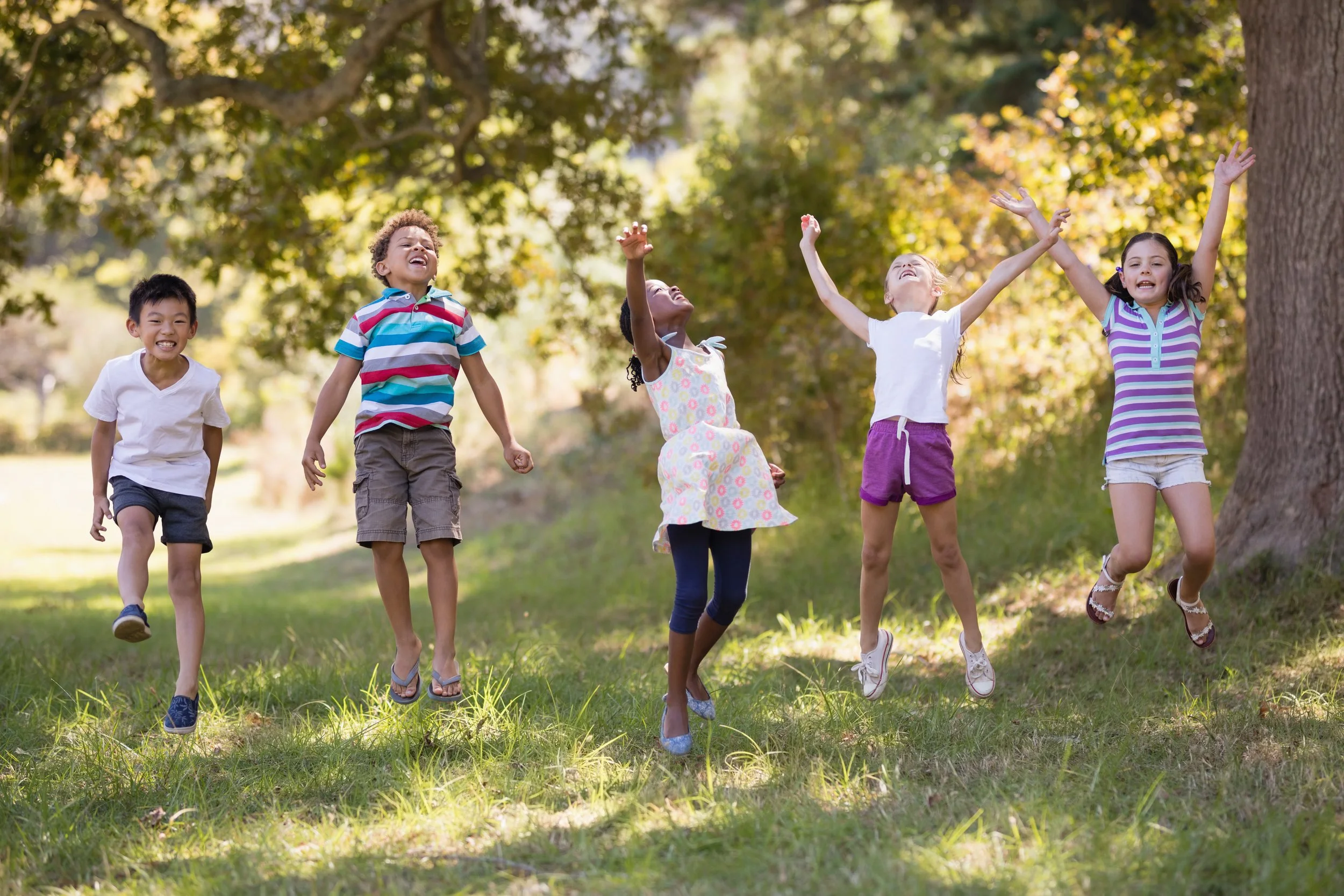 Group of five children running and playing outdoors in a park during daytime with trees and sunlight.