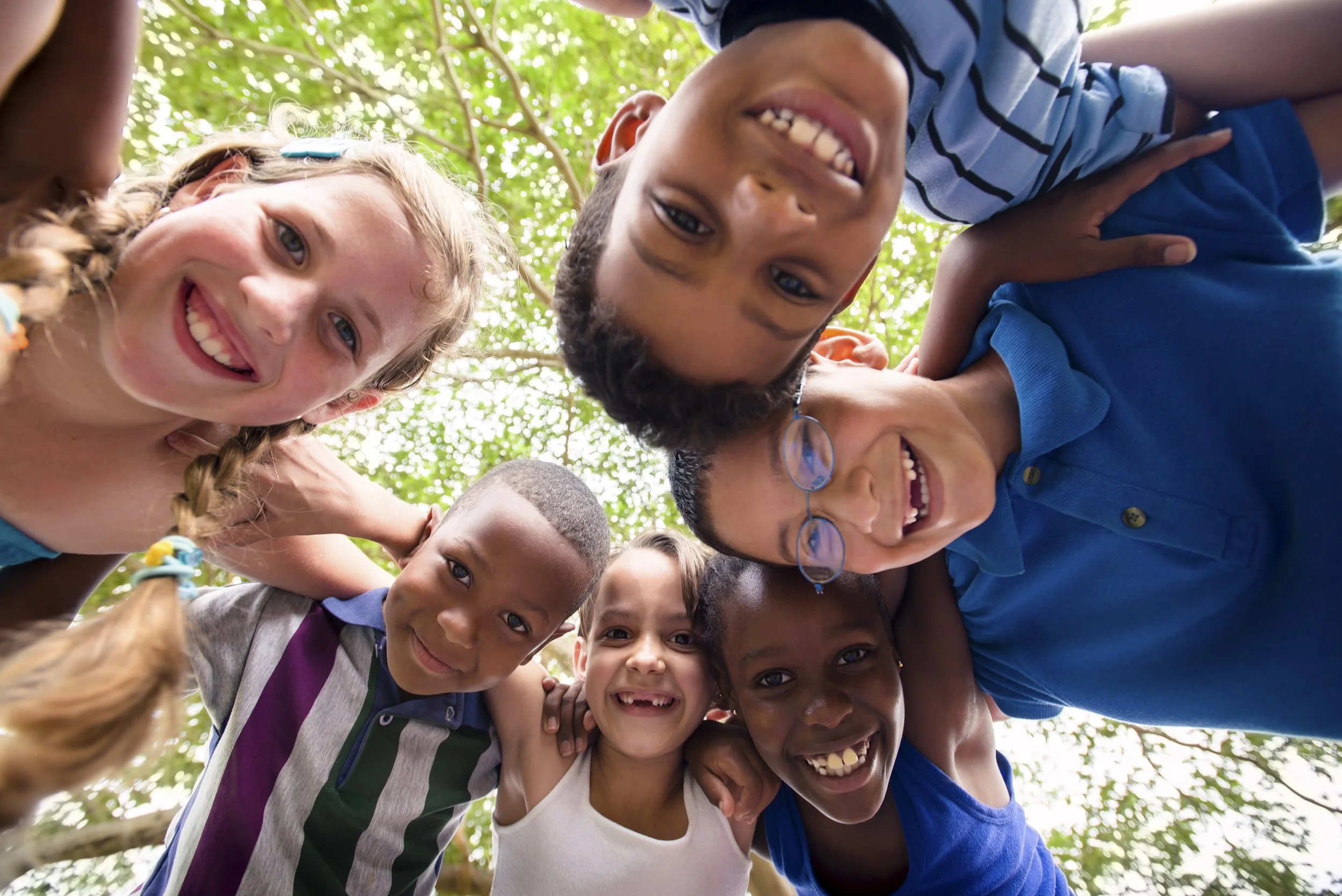 Group of pre-teen children smiling and huddling together outdoors, looking down at the camera, with green leaves and tree branches above.