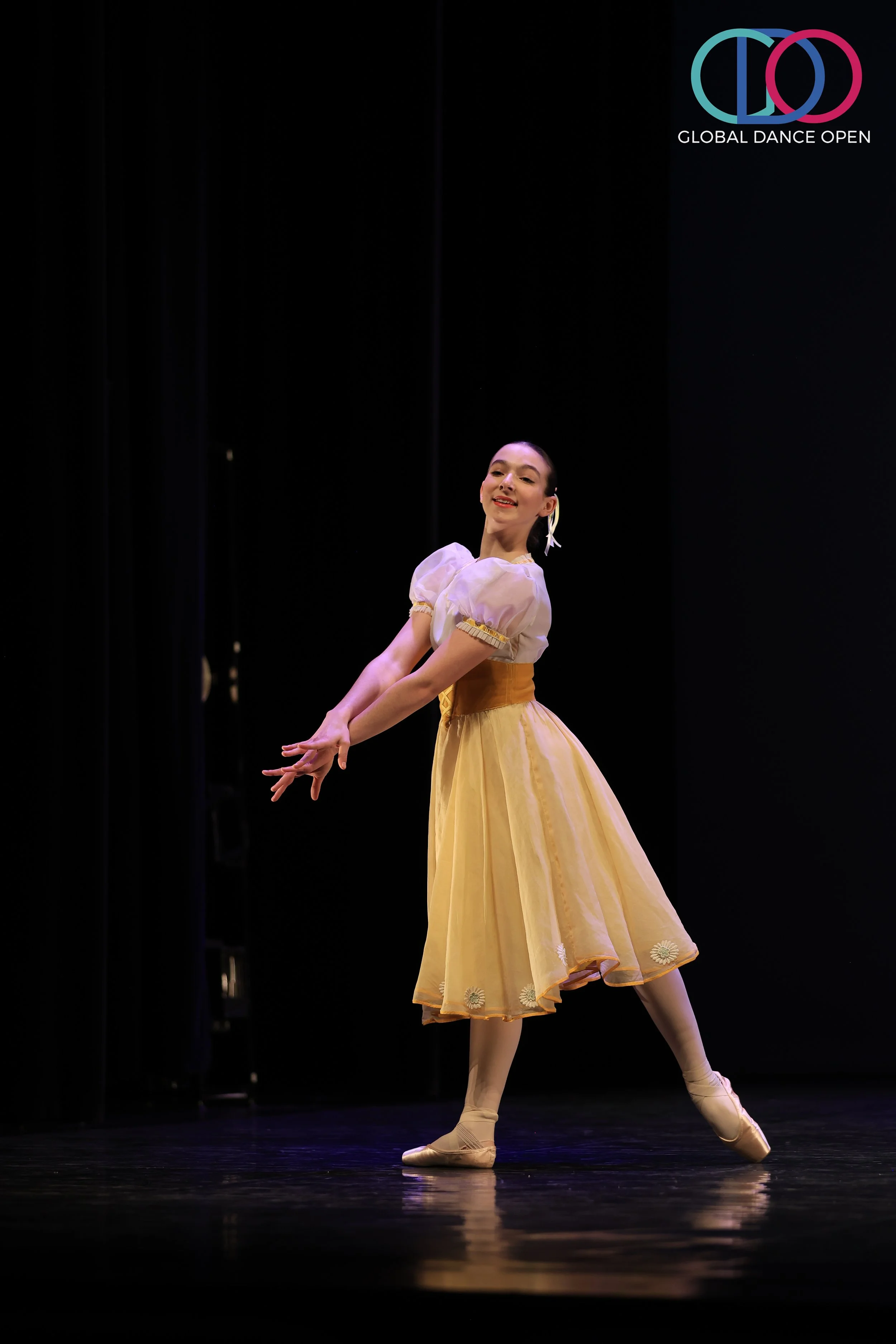 Een ballerina in een geel tutu danst op een donker podium. In de rechterbovenhoek het logo van de Global Dance Open.