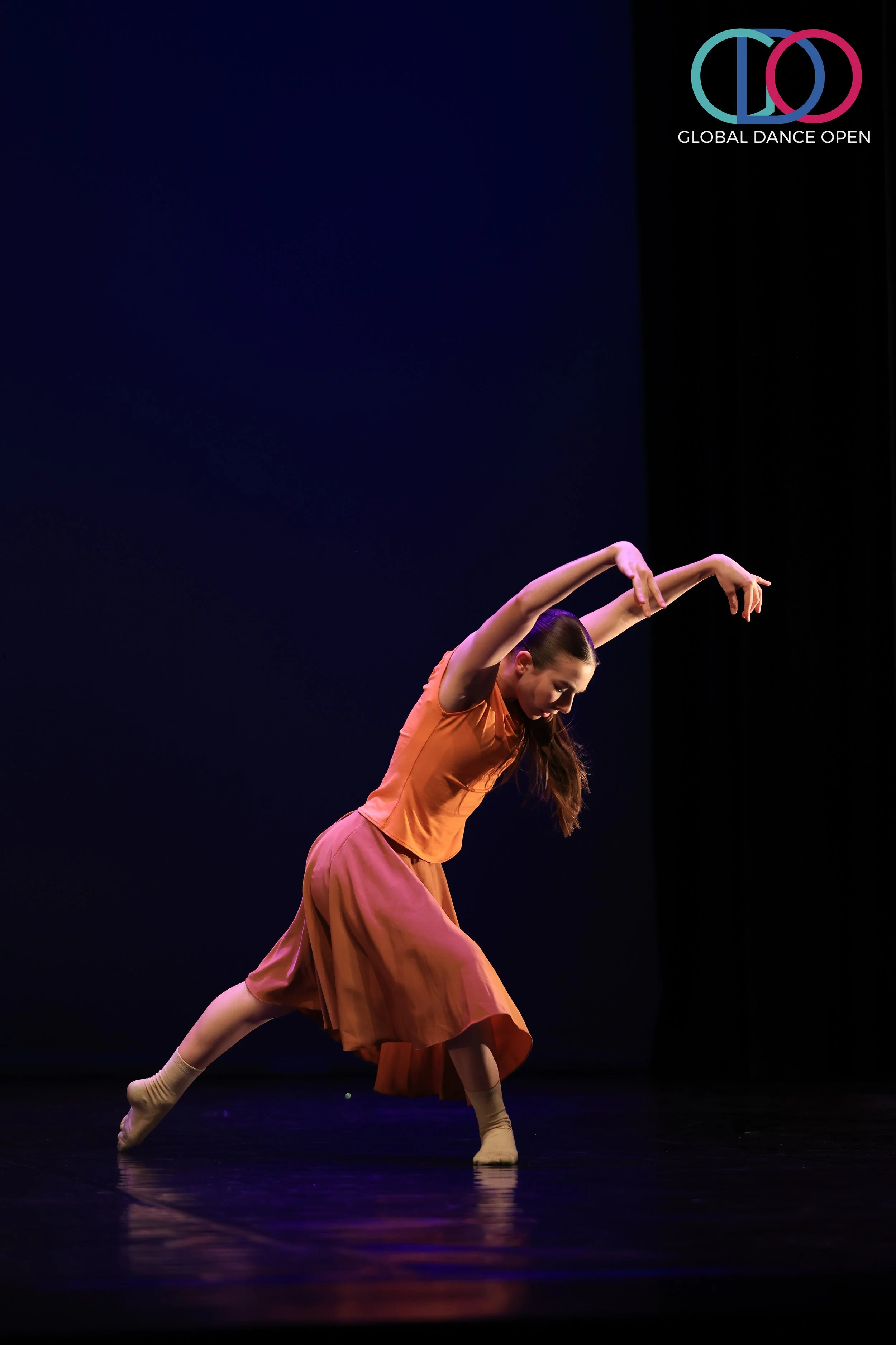 Dansende vrouw op een donkere podium, gekleed in een oranje shirt en roze rok, met armen omhoog en op een manier die dans bewegingen uitdrukt, bij de mondiale danscompetitie.