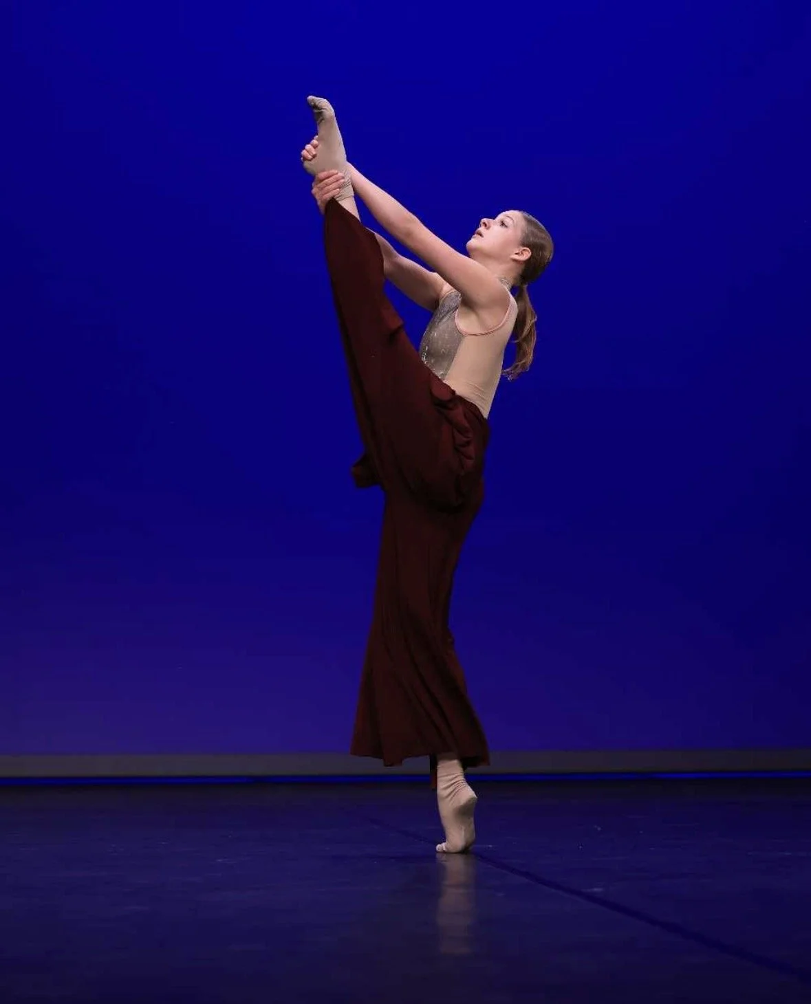 Een danseres voert een kielhouding uit op het podium tegen een blauwe achtergrond.