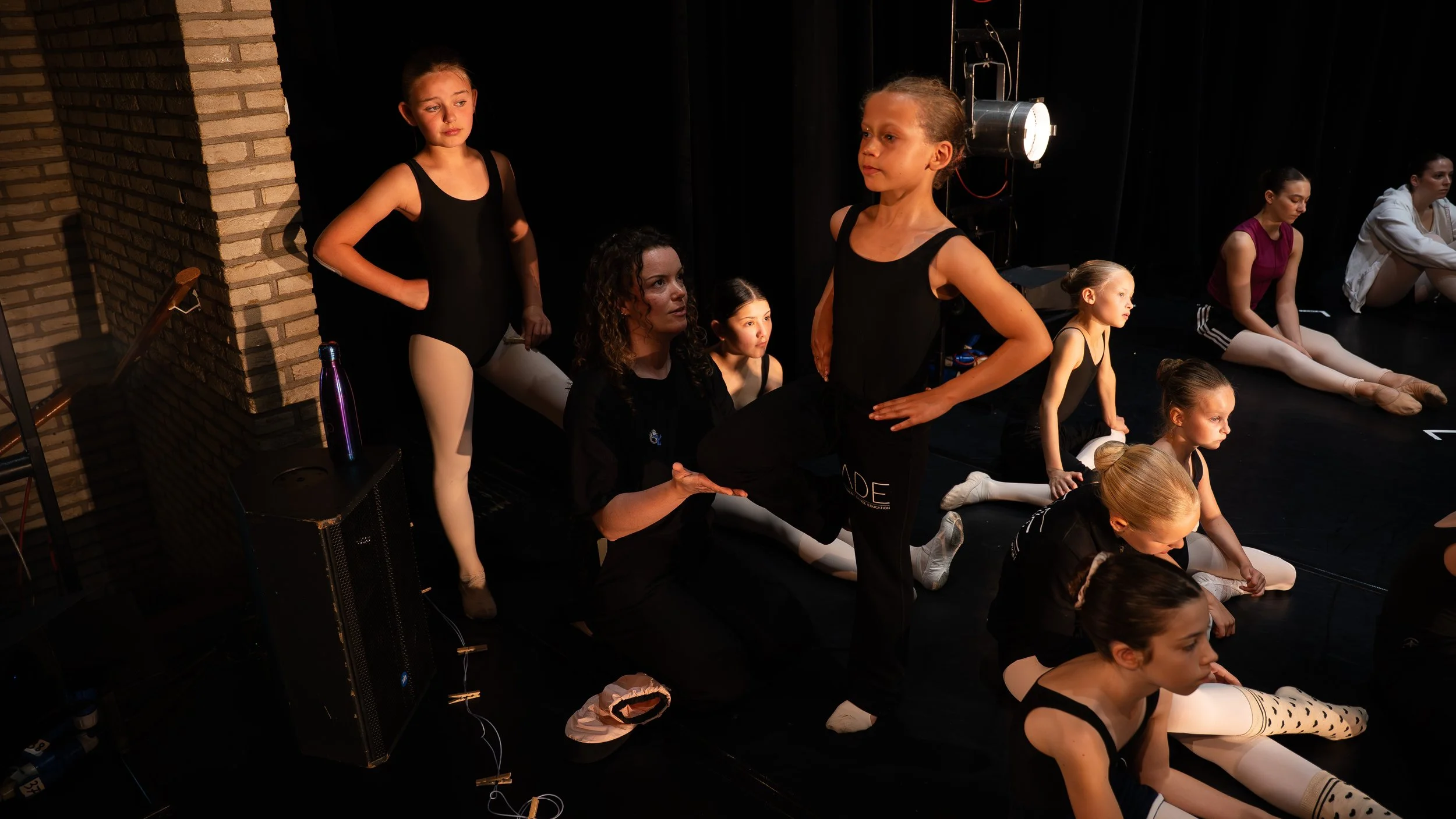 Balletdanseressen in een repetitie of voorstelling, sommige zitten of liggen op de vloer, andere staan in een zwarte toneelomgeving.