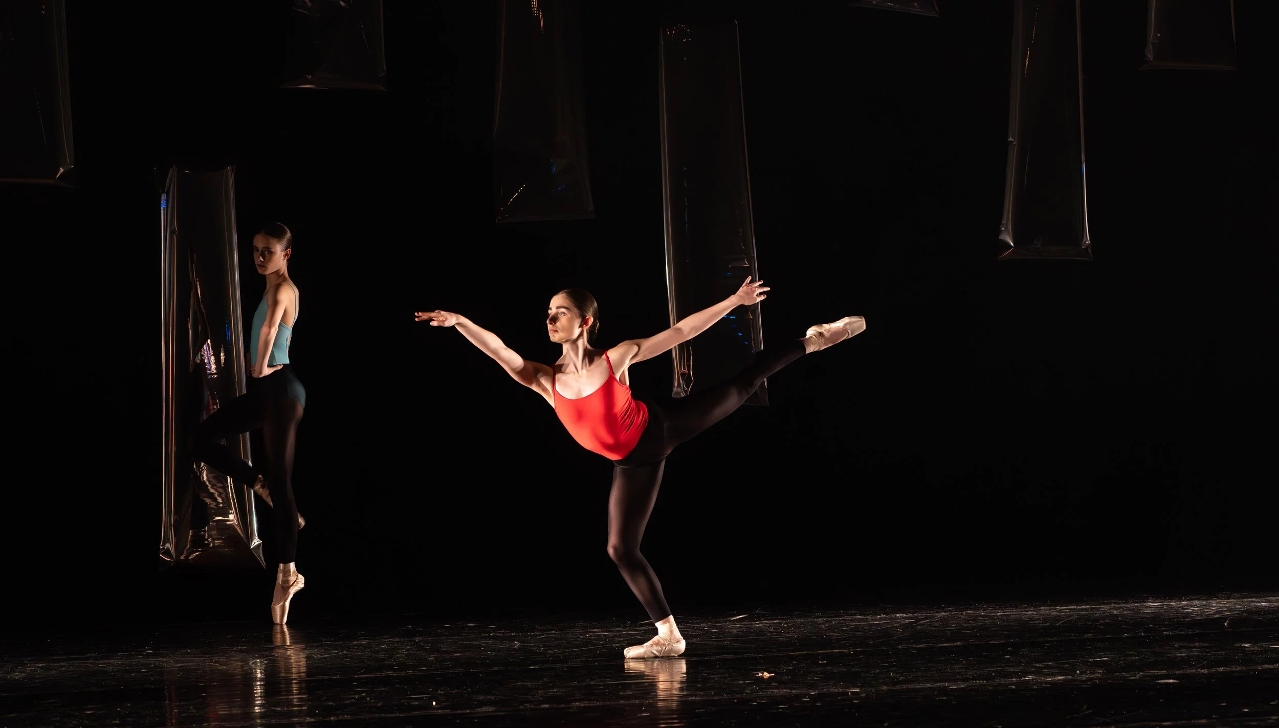 Twee balletdansers oefenen op een donkere achtergrond, één in een rode top en zwarte broek, de andere in een blauwe top en zwarte broek, tijdens de repetitie.