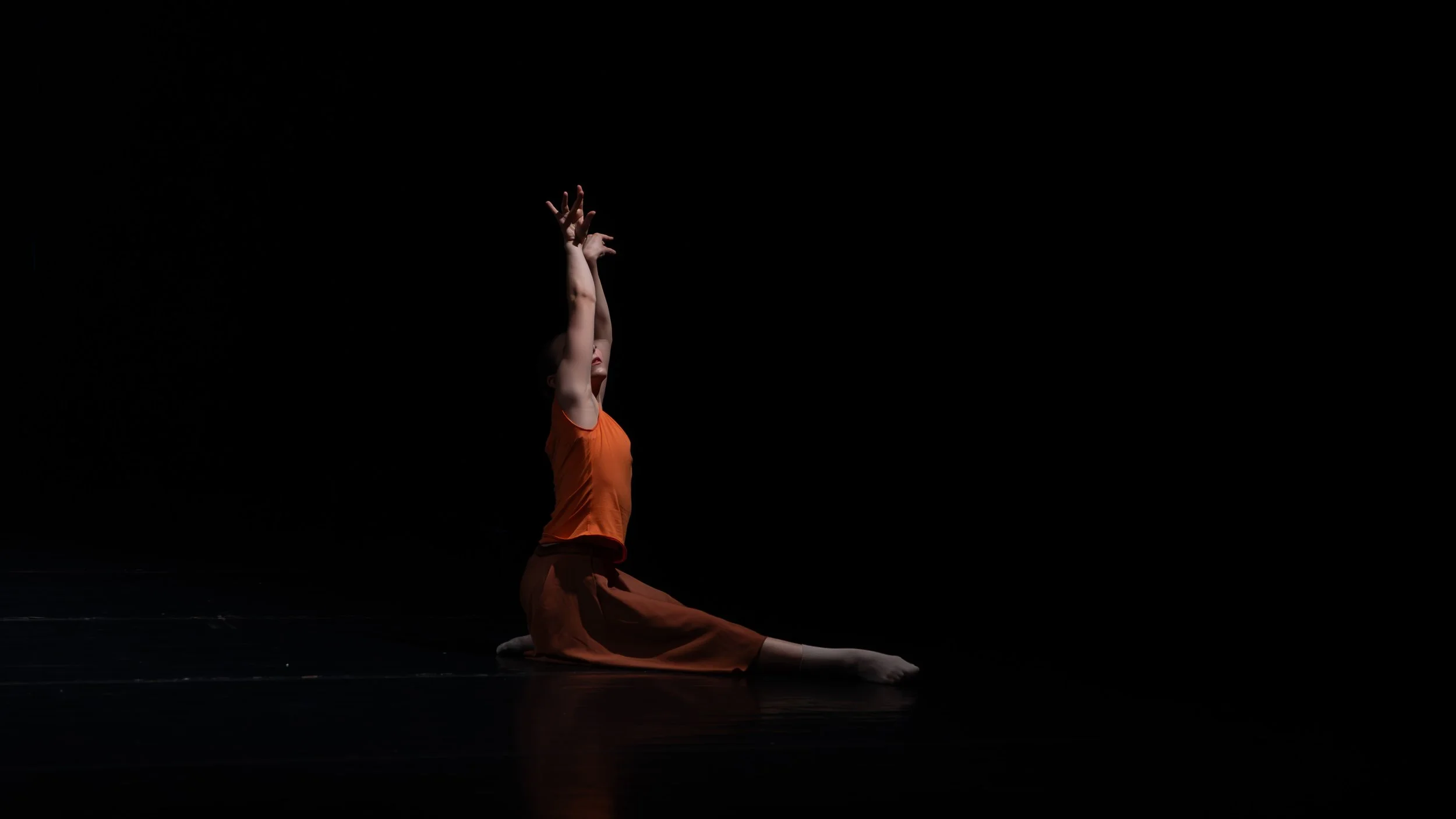 Een danseres in een oranje outfit voert een posteriore bocht uit op een donkere achtergrond.