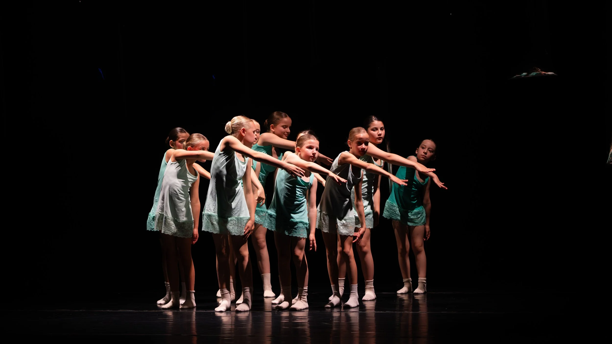 Tien jonge meisjes in witte en turquoise dansoutfits dansen op een donker podium.