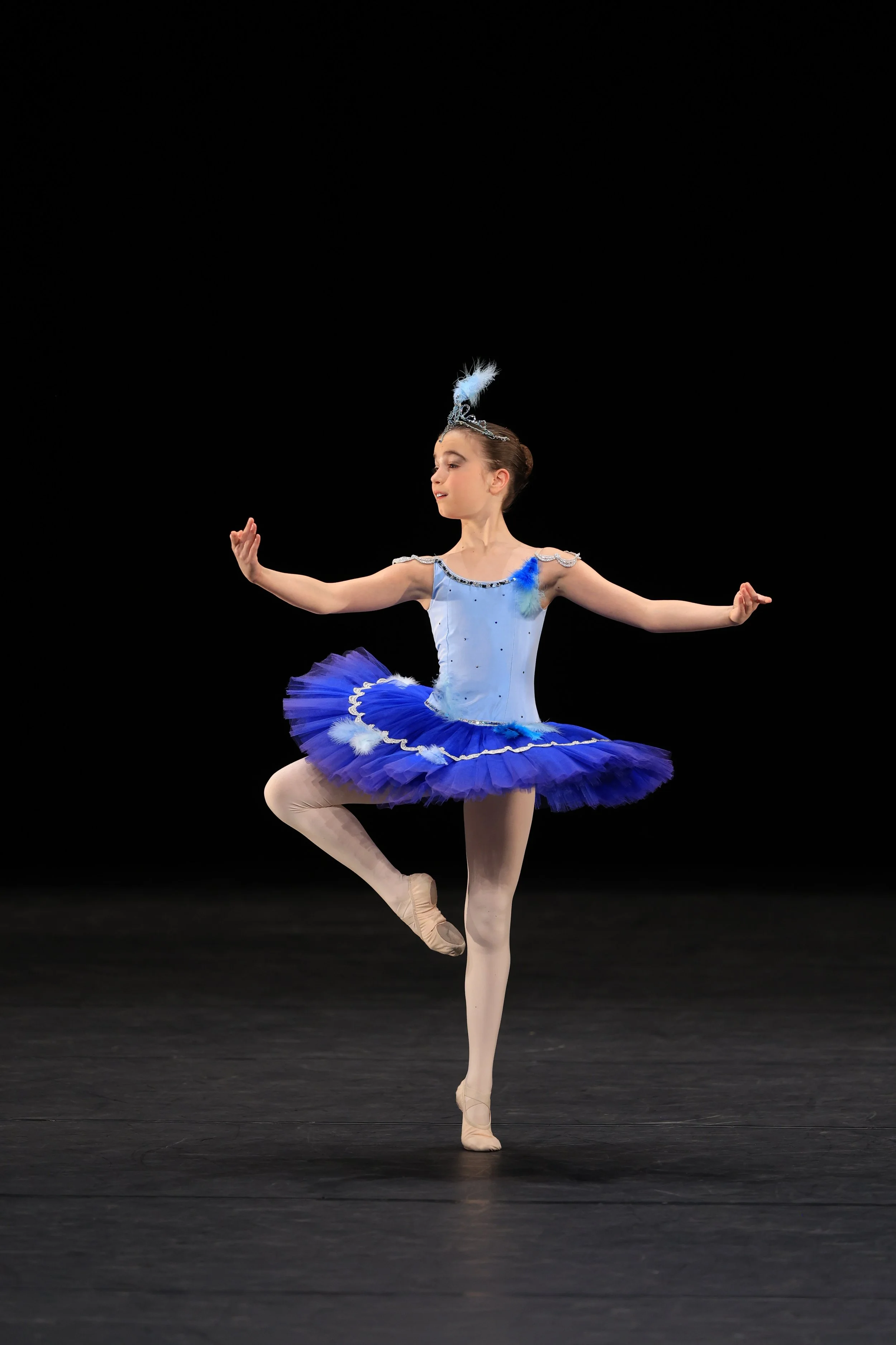 Een jonge ballerina in een blauwe tutu voert een danspositie uit op een donkere podiumvloer.