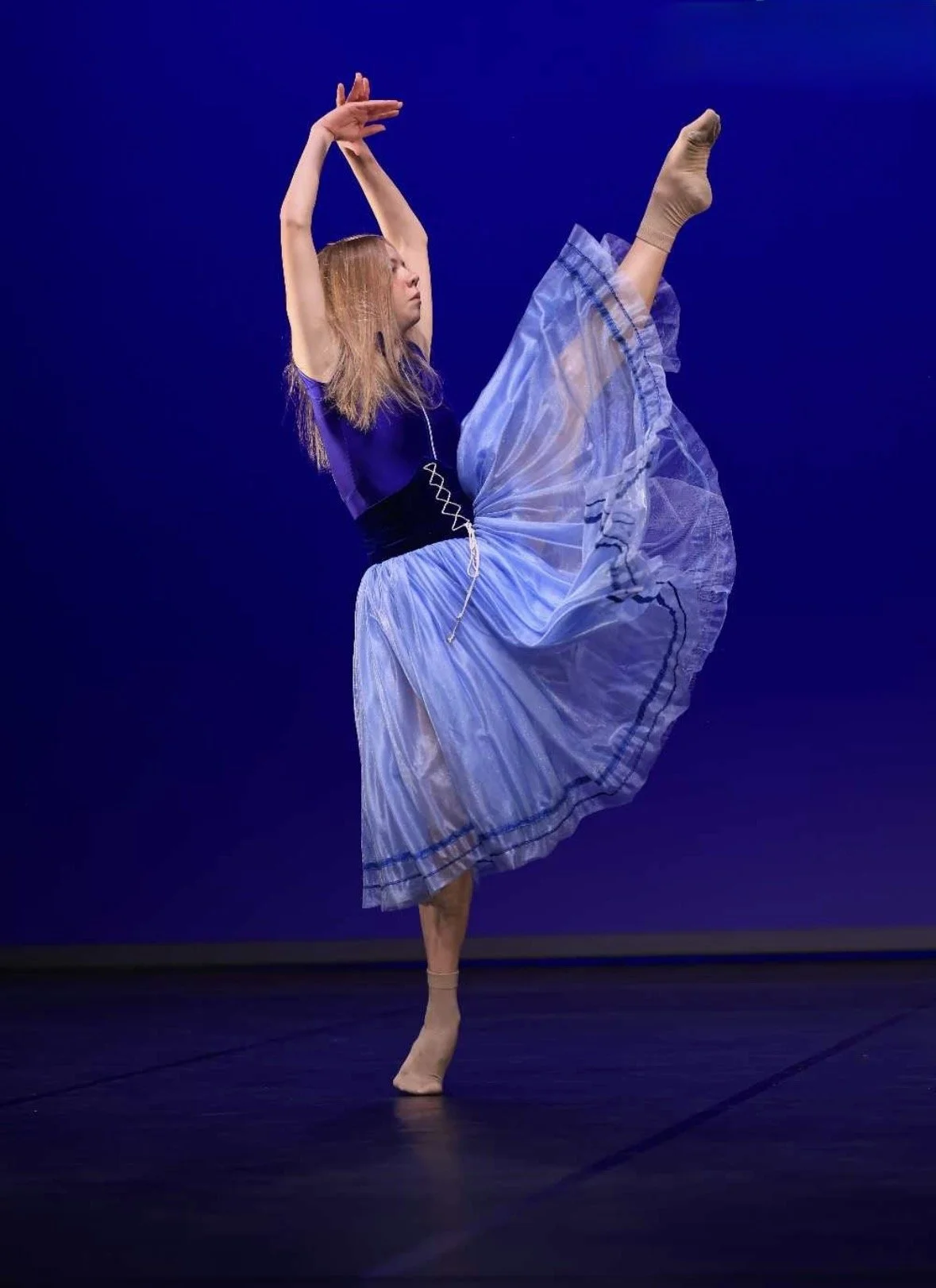 Balletdanseres in een blauwe tutu voert een sprong uit op een donkere podium, met een blauwe achtergrond.