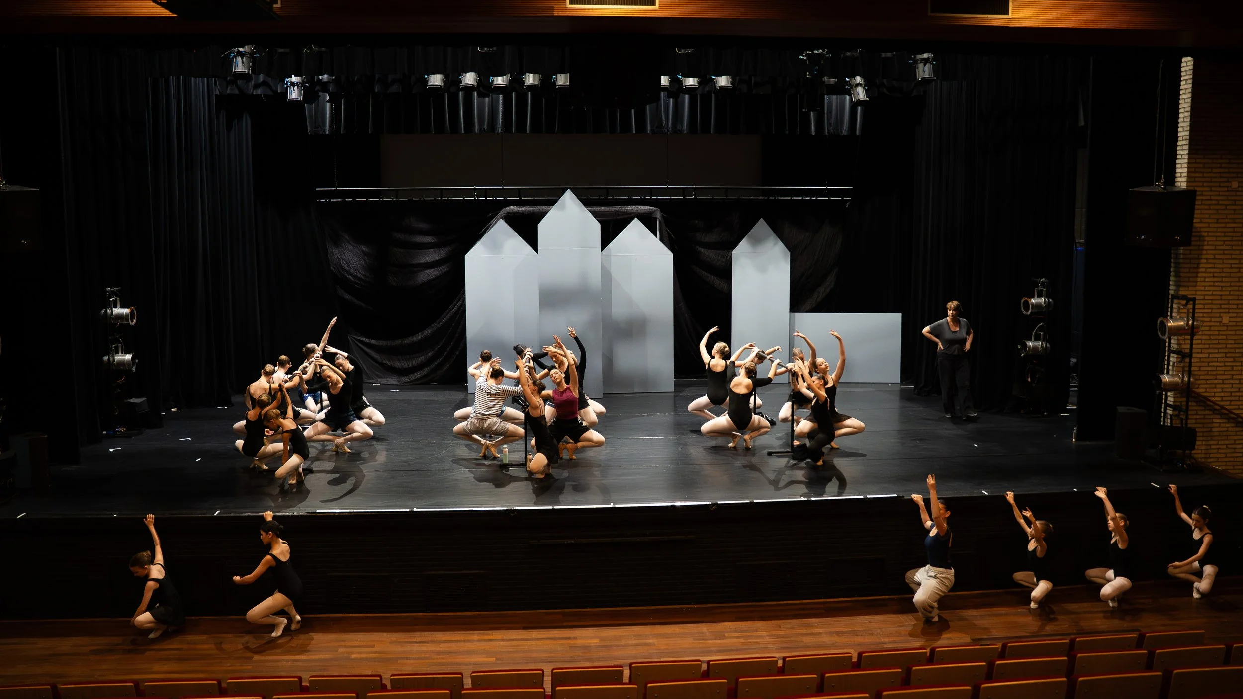 Balletdansers oefenen op een podium met zwart gordijn en witte decorstukken, instructeur waakt toe terwijl de dansers in verschillende posities zitten en bewegen.