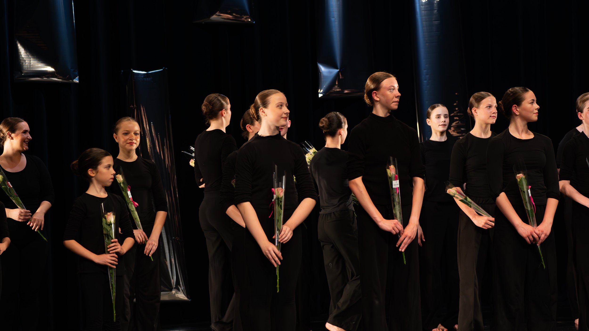 Vrouwen en meisjes in zwarte kleding die bloemen vasthouden staan op een toneel, mogelijk na een podiumoptreden of ceremonie.