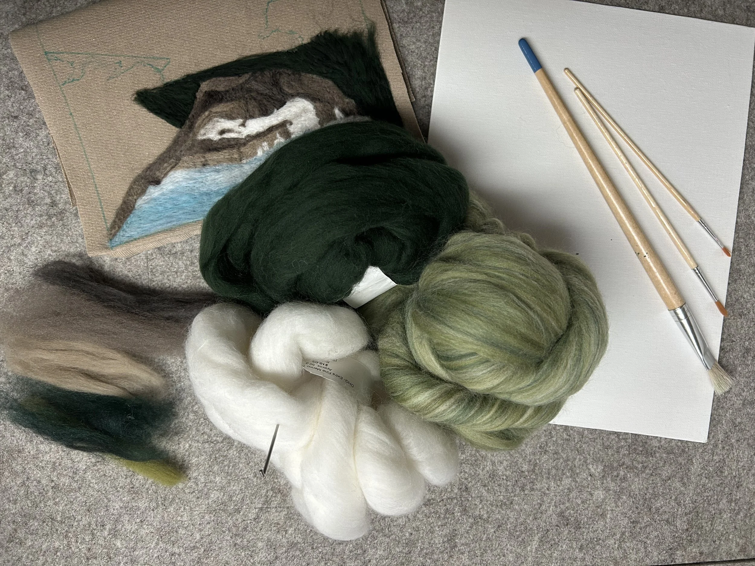 Balls of yarn in various colors including green, white, black, and a multi-colored skein, placed on a gray textured surface with a blank white piece of paper and three crochet hooks nearby.