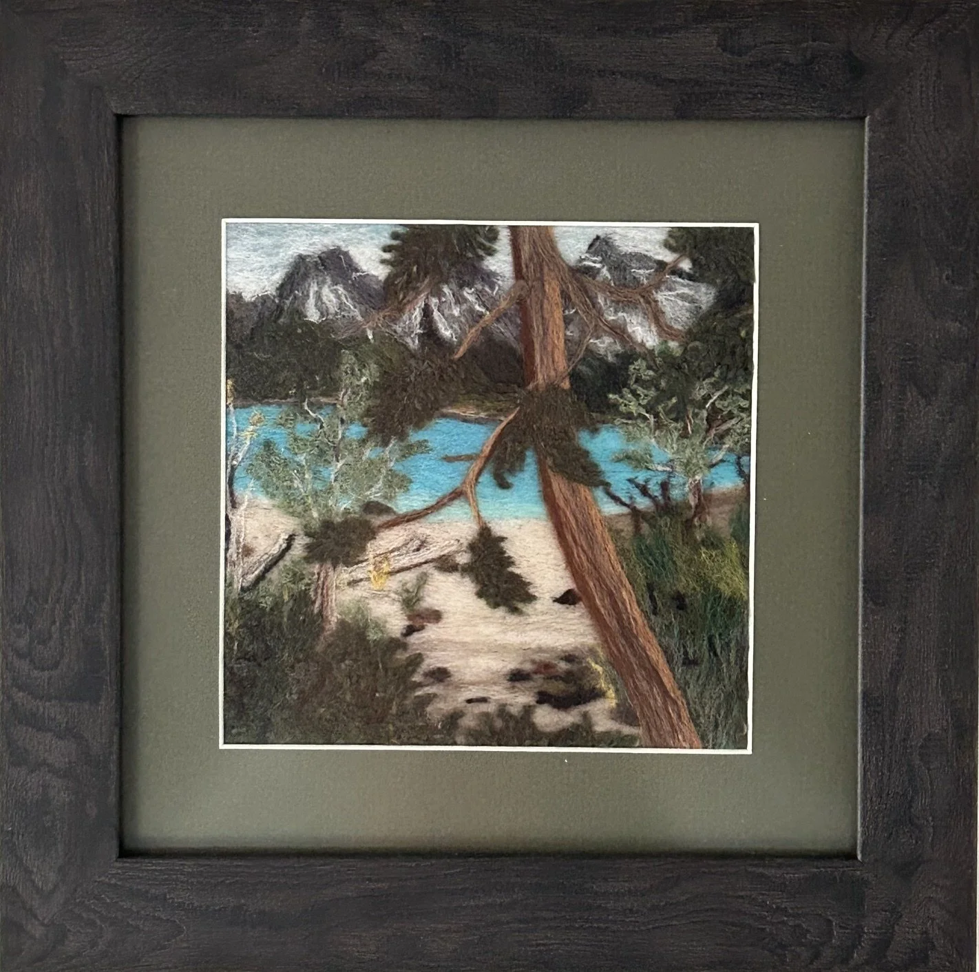 .Needle felted landscape of mountains, and a sandy beach on a blue lake, viewed from behind a pine tree.