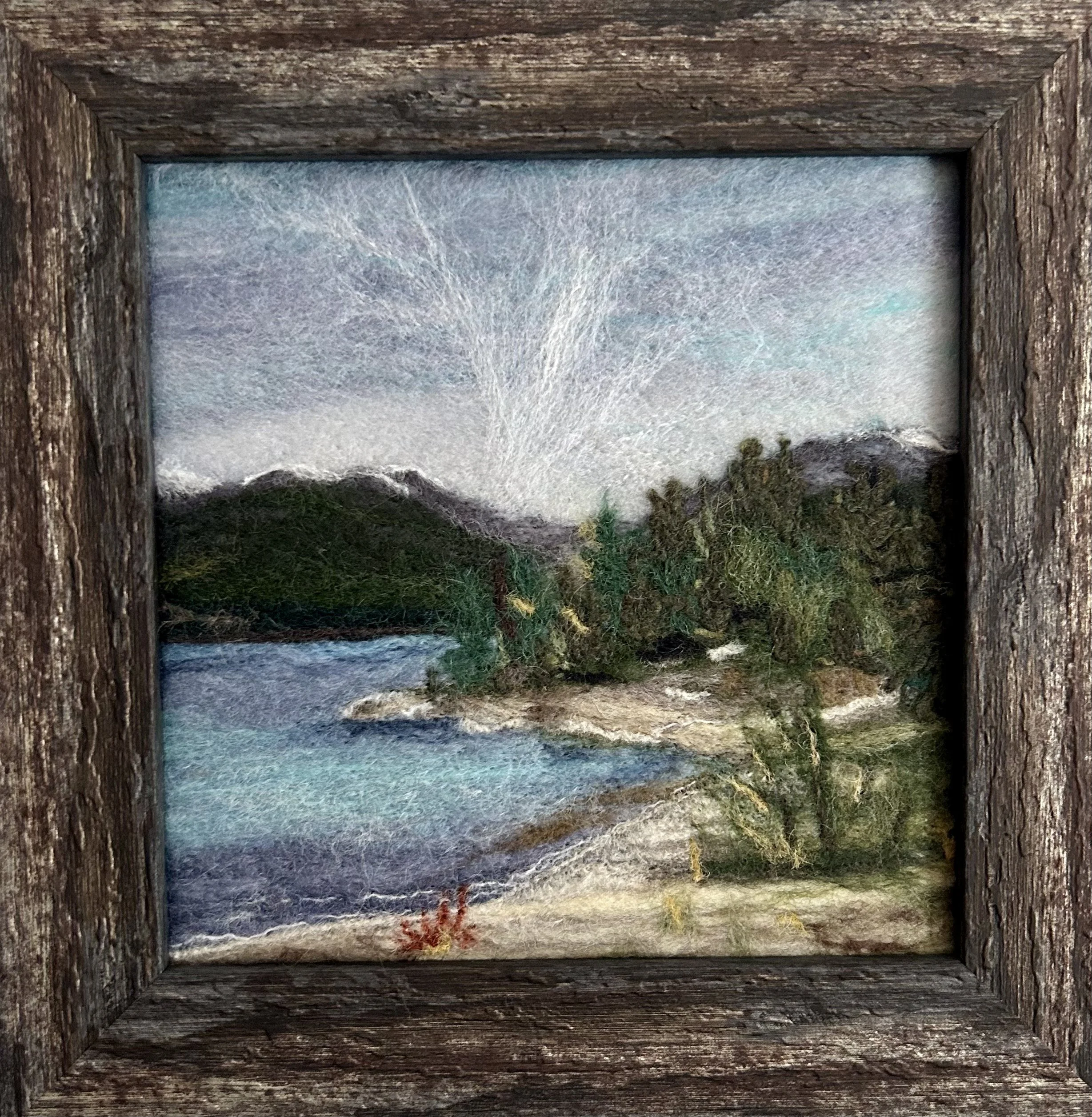 A framed artwork featuring a mountain landscape with trees, water, and sky, seen through a rustic wooden frame.