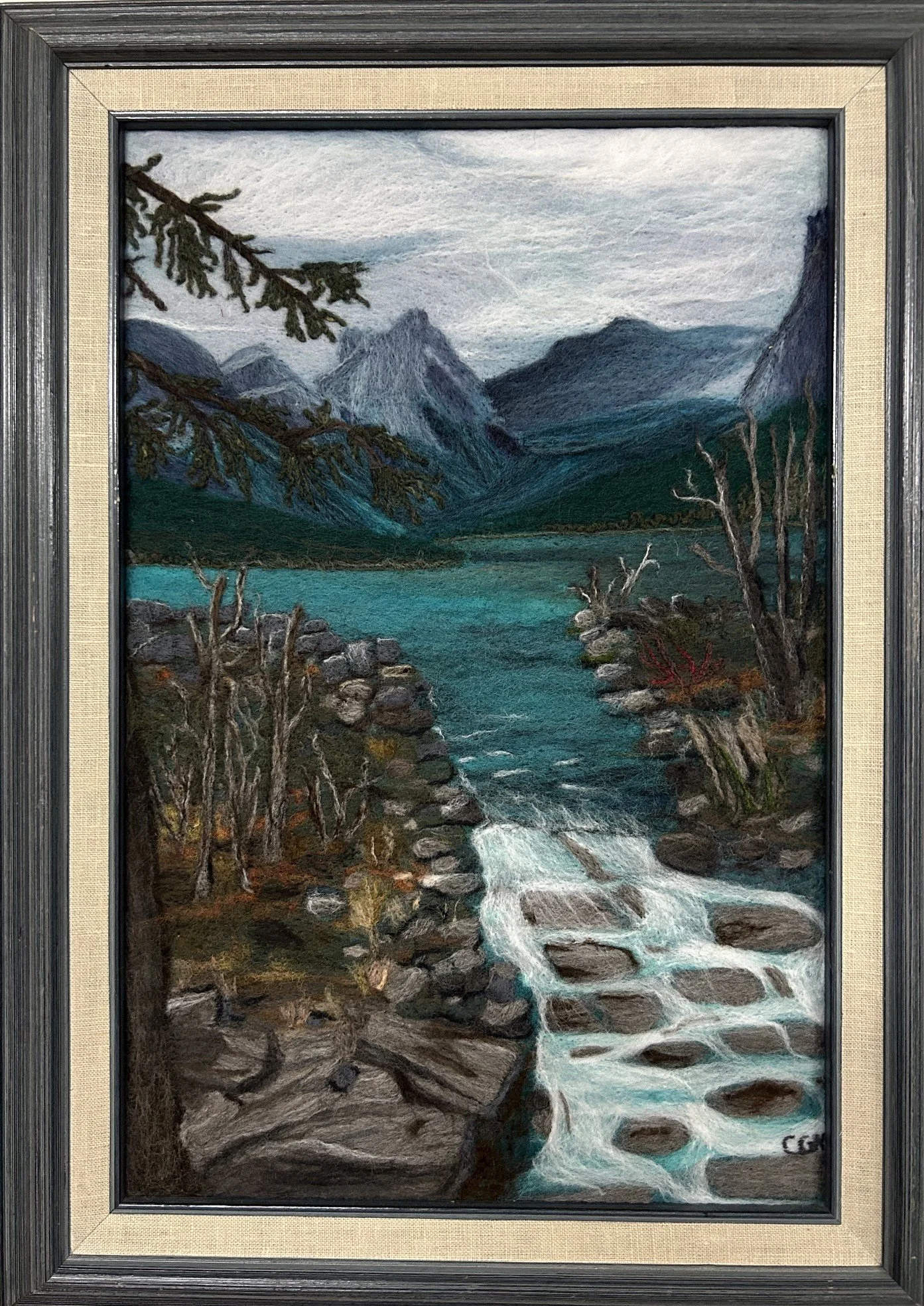 A felted landscape painting of mountains with a river flowing down to a small rapid creek with a rocky terrain and driftwood, with a cloudy sky overhead.