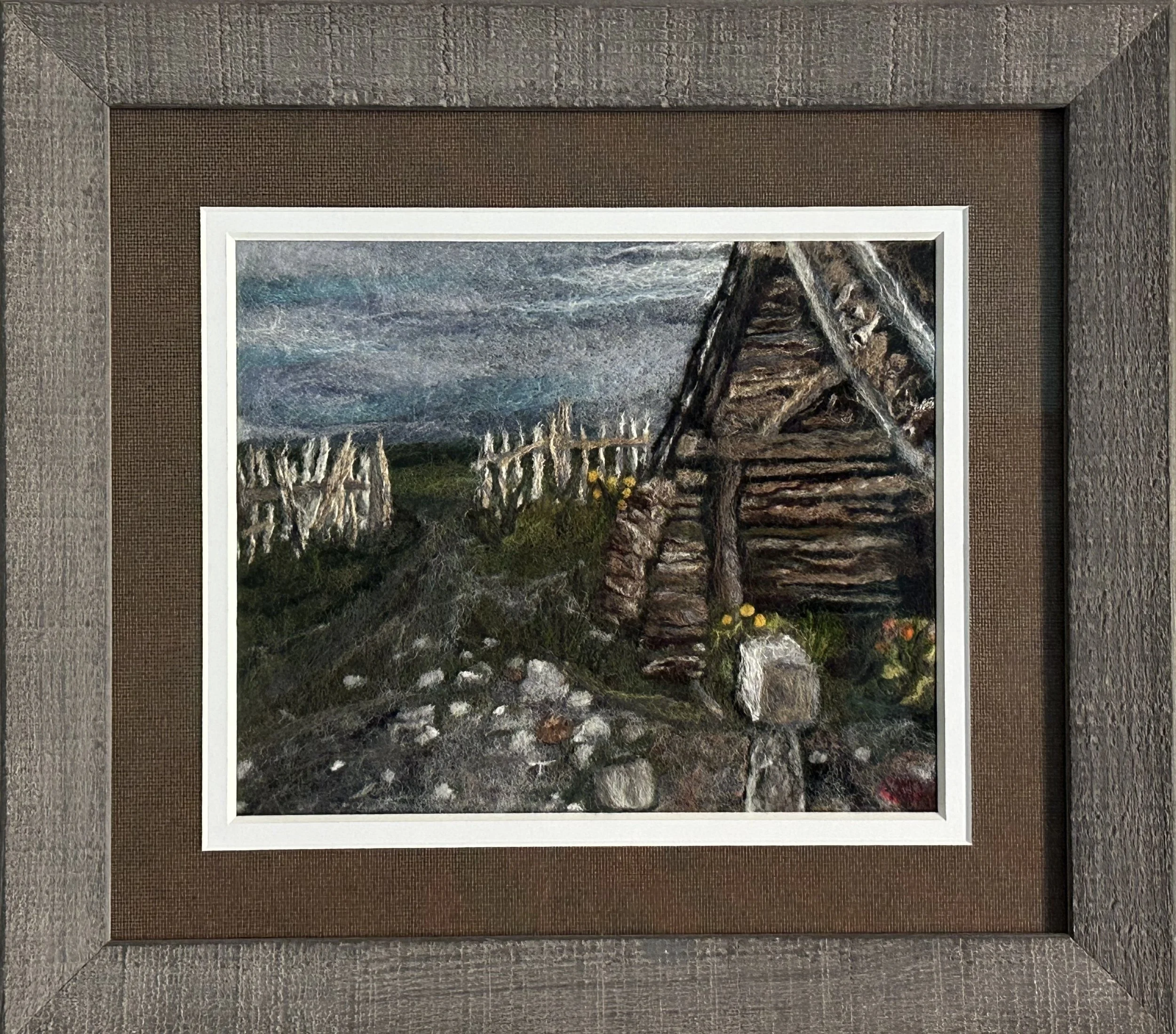 A felted depiction of an old weathered Woodshed and fence on a windblown prairie with a stormy sky.